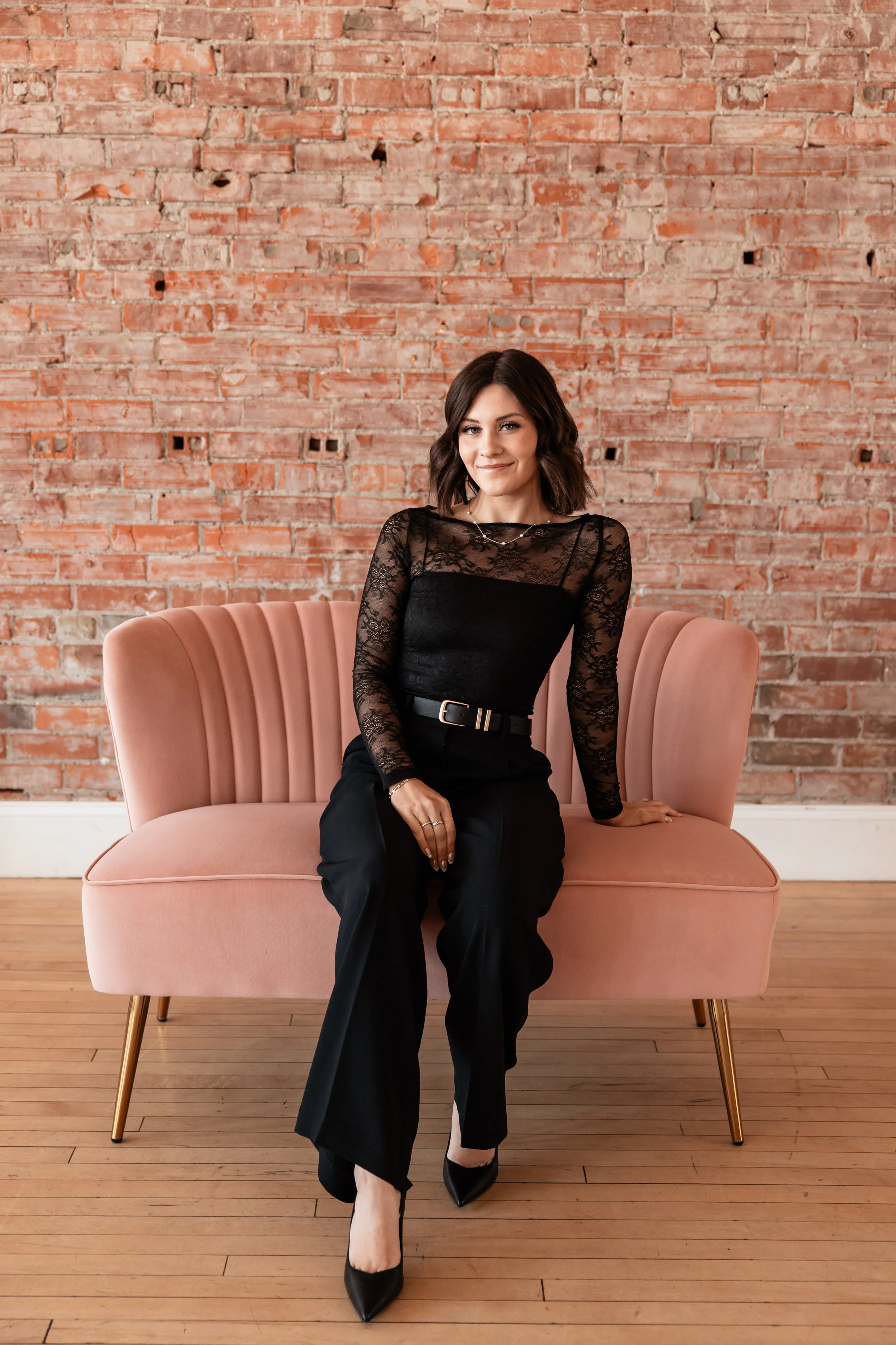 A woman with shoulder-length dark hair wearing a black lace top and black pants, sitting on a pink velvet sofa with a pink brick wall in the background.