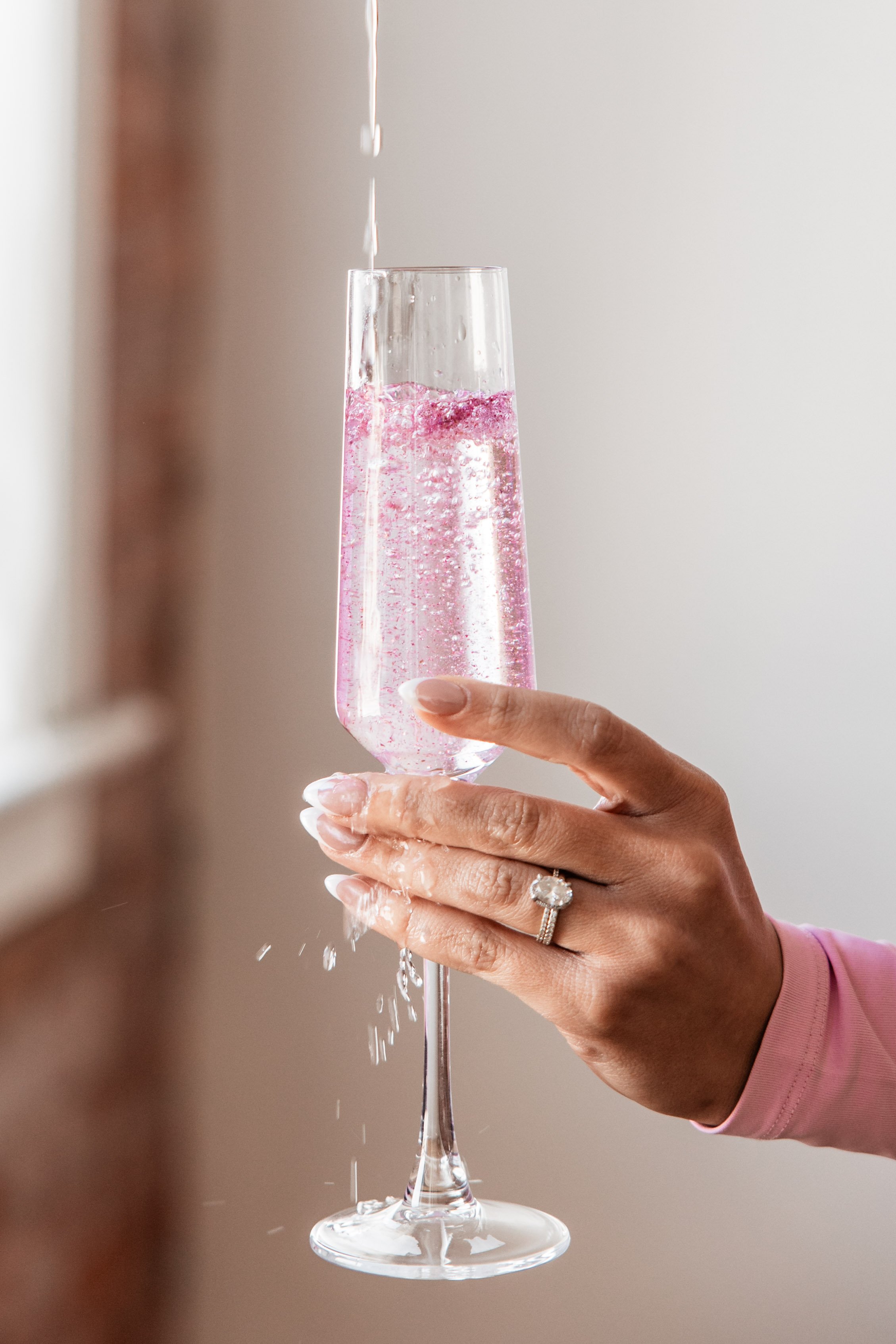 Hand holding a tall, clear champagne glass filled with pink sparkling wine, with some bubbles visible. The hand is wearing a wedding ring and a pink sleeve, and water is stream from above into the glass, causing some splashing.