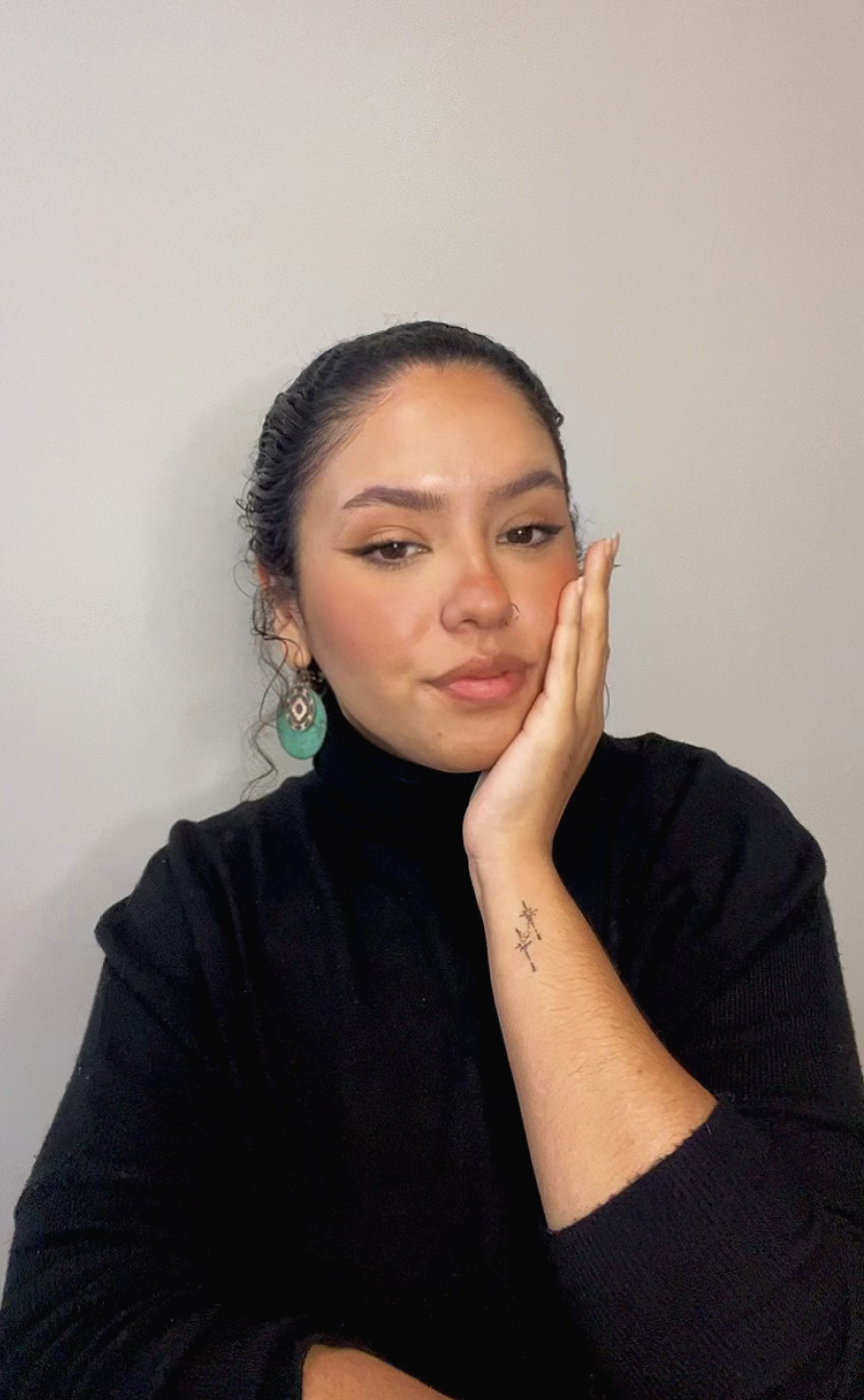 A woman with her hand resting on her cheek, wearing a black top, earrings, and has a tattoo on her forearm, posing against a plain light-colored wall.