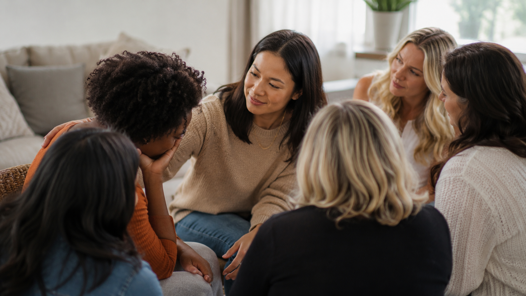 Group of women sitting together indoors in a living room, one woman comforting another woman who appears to be upset, while others watch and listen.