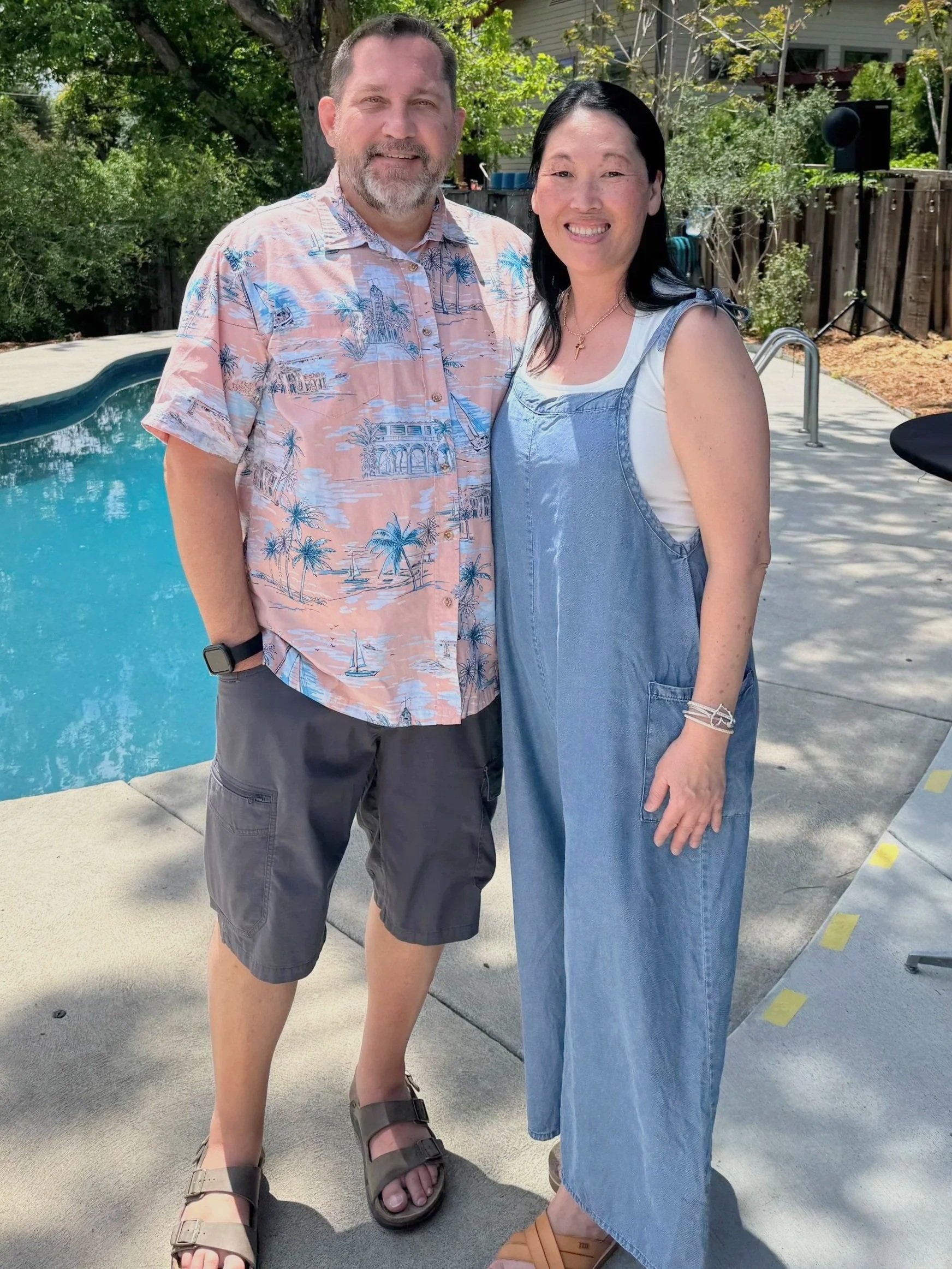 A man and a woman standing outdoors by a swimming pool, smiling at the camera. The man is wearing a pastel-colored tropical shirt, gray shorts, sandals, and a smartwatch. The woman is dressed in a white top with a blue denim overall dress, wearing a cross necklace, and sandals. There are trees, a wooden fence, and a house in the background.