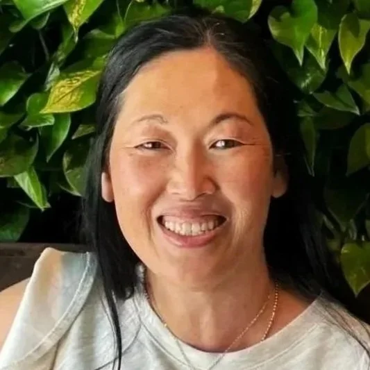 A smiling woman with long black hair, wearing a light-colored top, in front of green leafy plants.