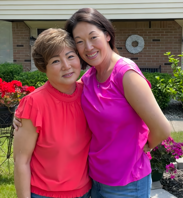Two women standing outside in front of a house, smiling and hugging each other, with colorful flowers and green bushes in the background.