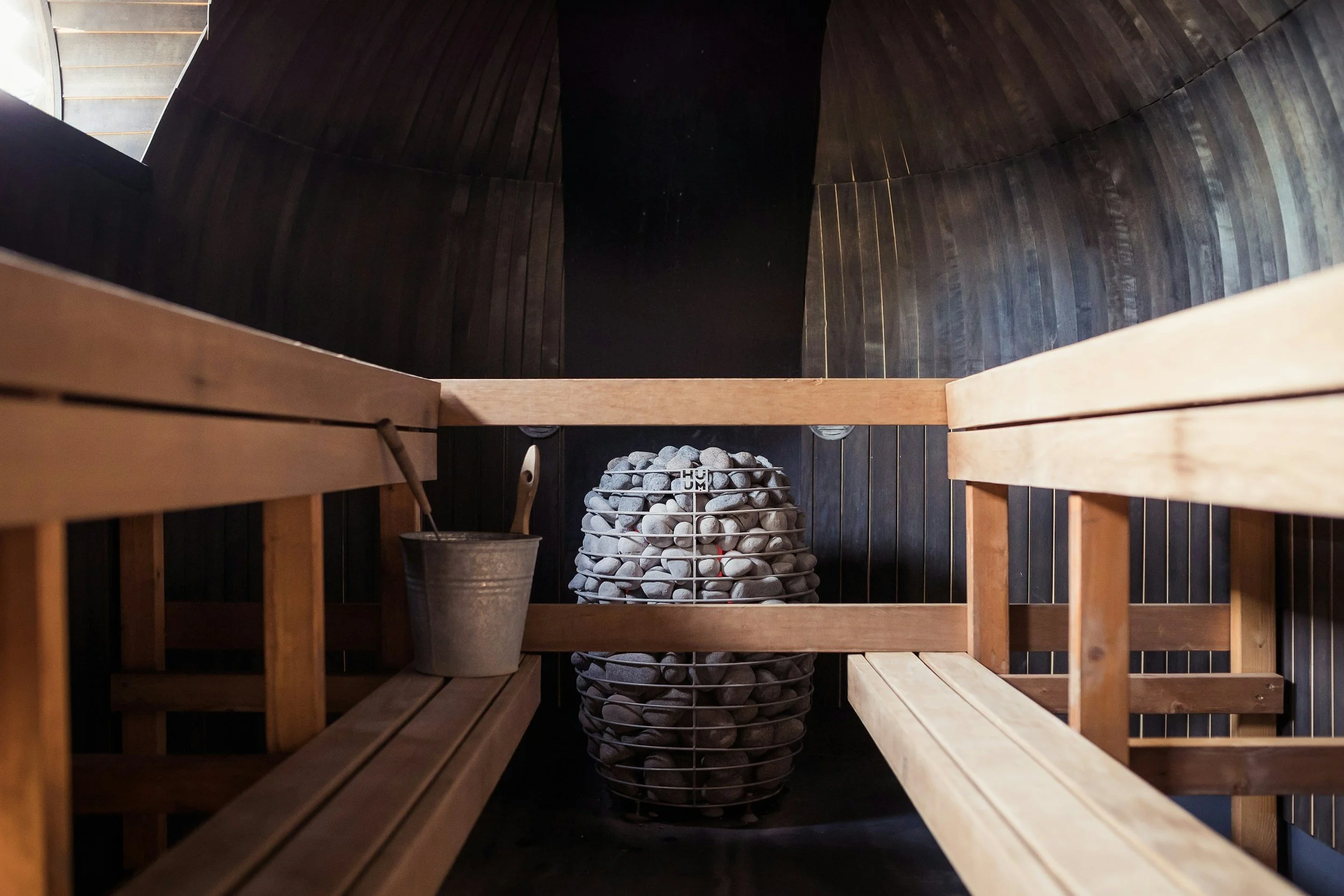Inside a sauna room, view from above showing wooden benches, a bucket of water with a ladle, and a heater filled with stones.