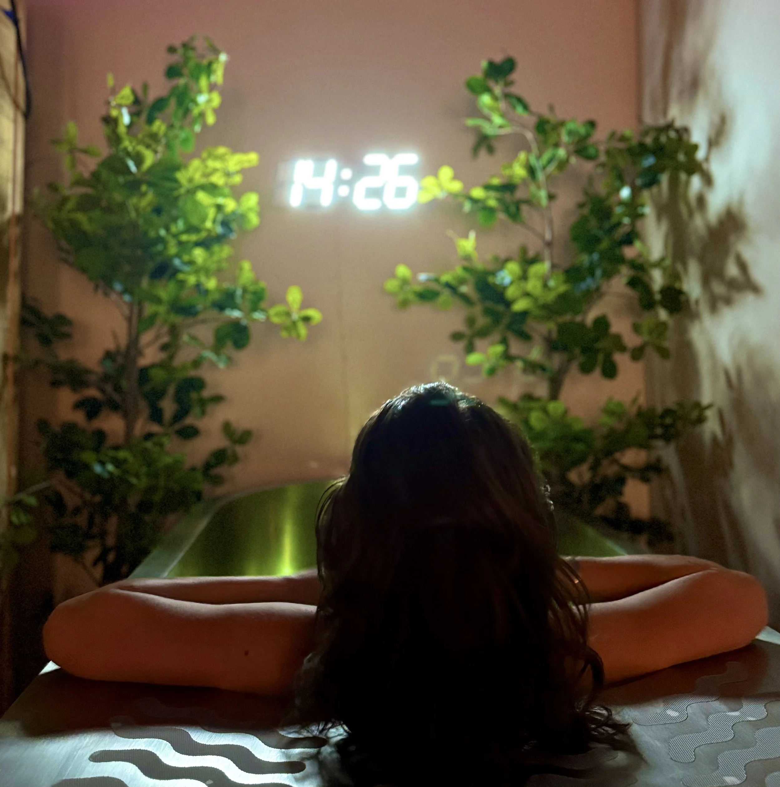 A person with long brown hair resting on a table with their arms extended, facing away from the camera. The background features two large green plants and a digital clock on the wall displaying 14:26.