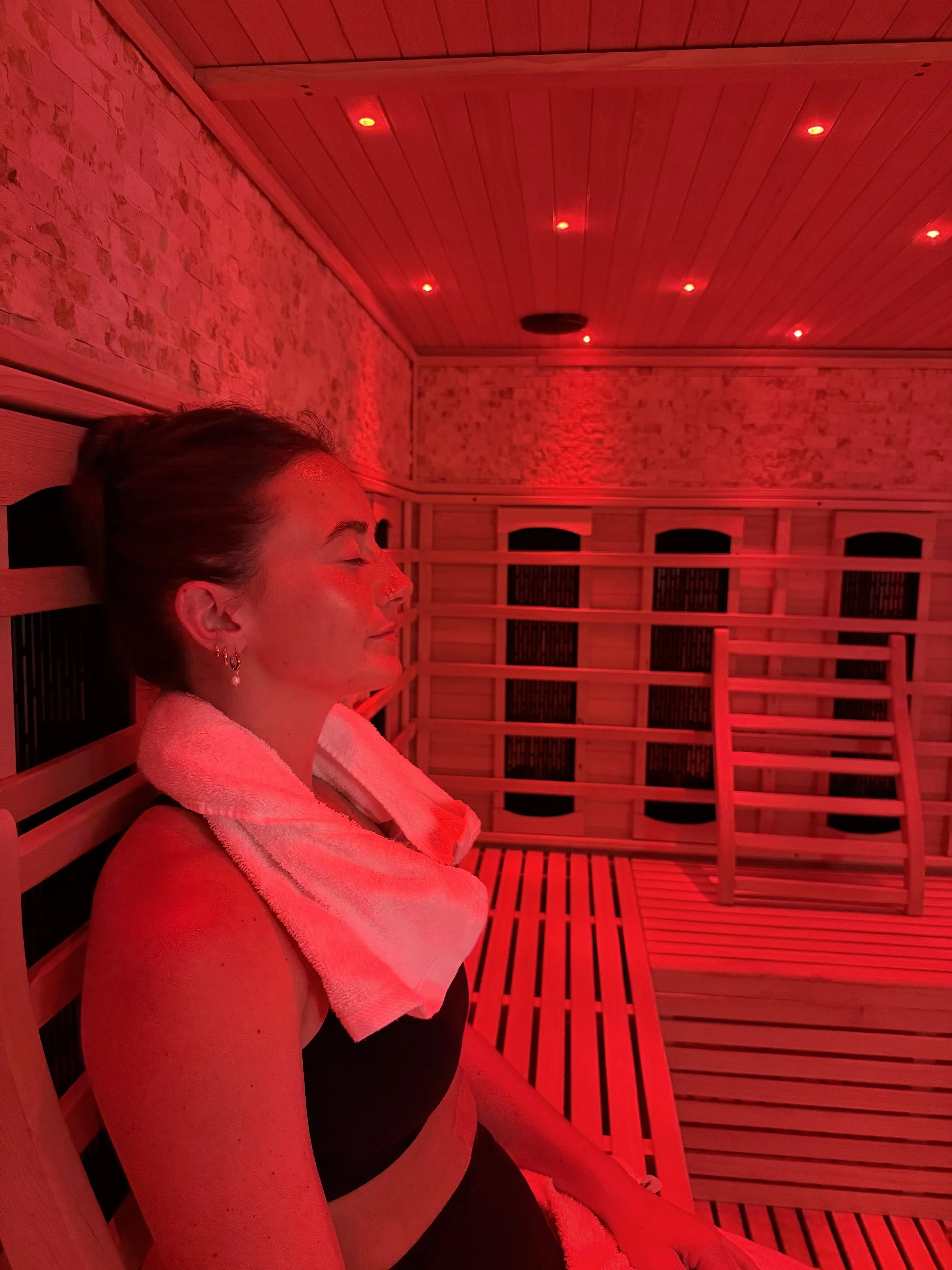 A woman is relaxing inside a sauna room illuminated by red light. She is sitting with her eyes closed, wearing a black top and has a towel around her neck. Wooden benches and walls are visible in the room.