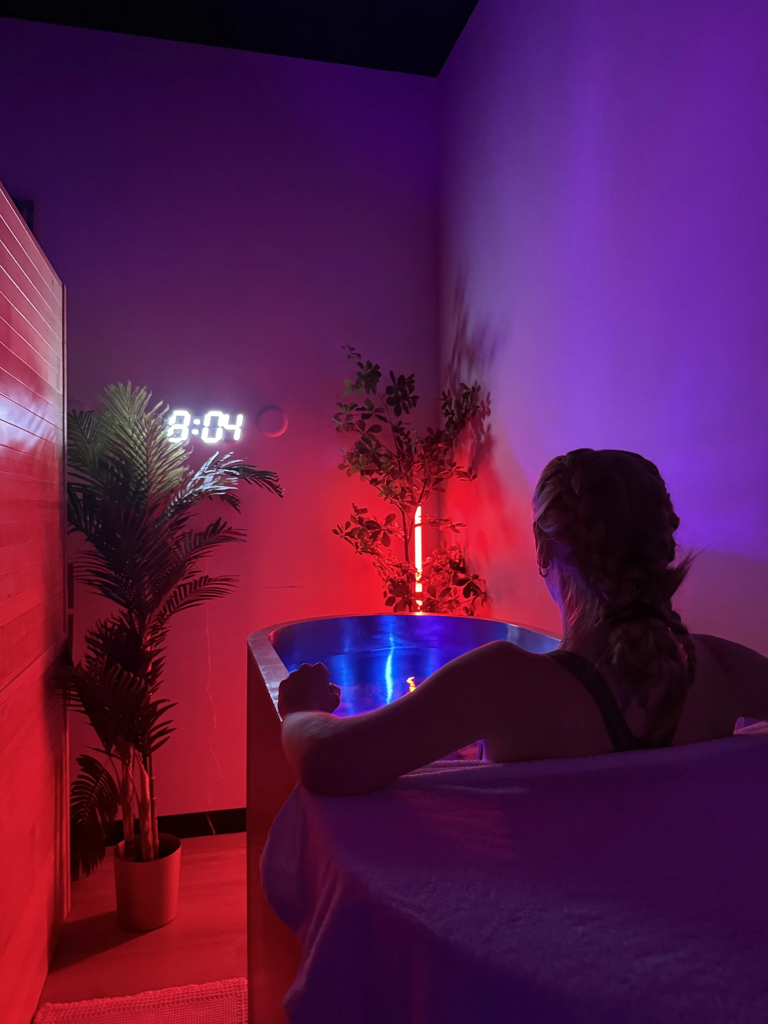 A woman relaxing in a bathtub illuminated by red and purple neon lighting, with plants in the background and a digital clock showing 9:04.