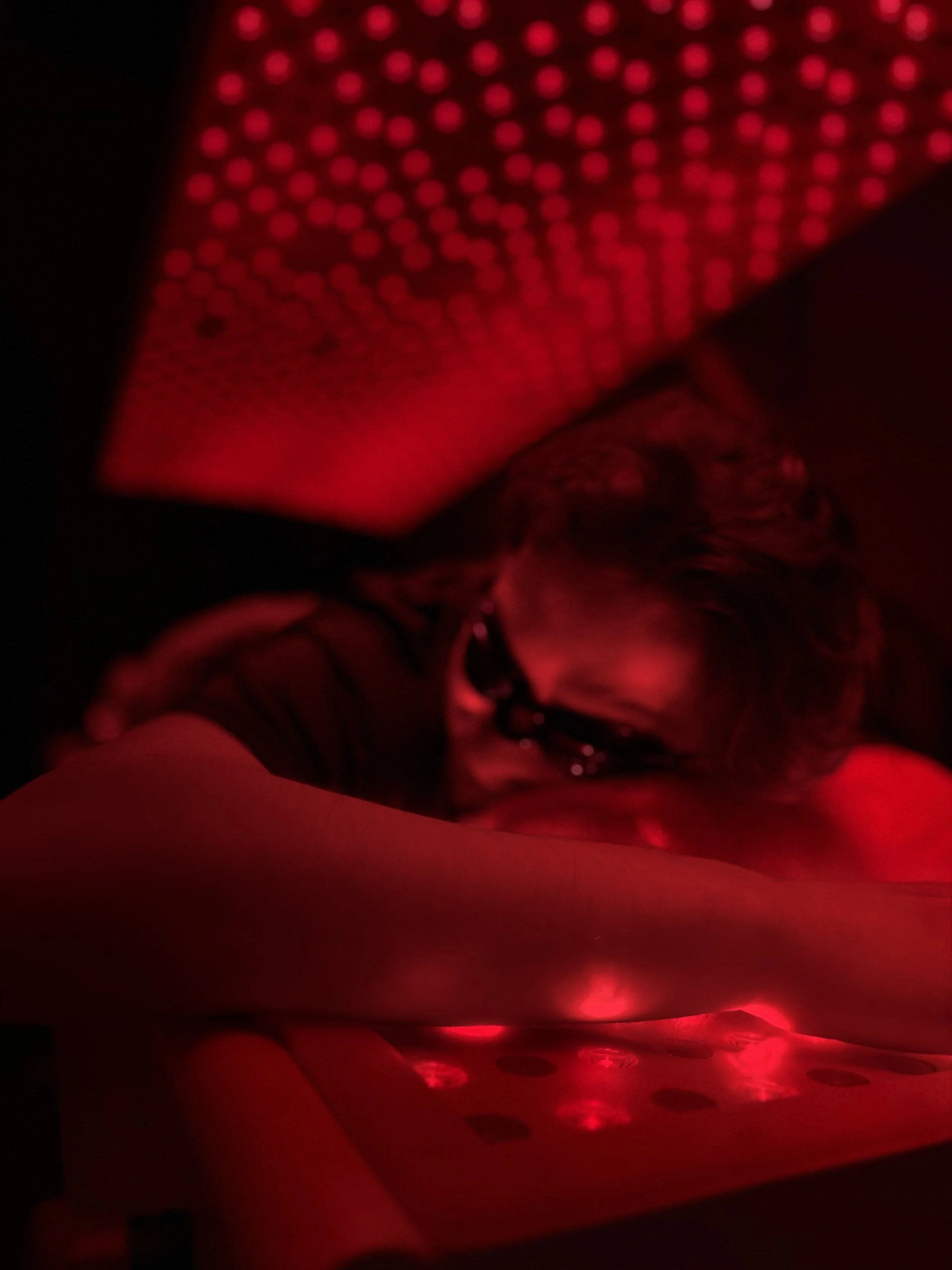 A person wearing glasses with curly hair, lying down with their head resting on a surface, illuminated by red LED lights, with a patterned light display on the ceiling above.