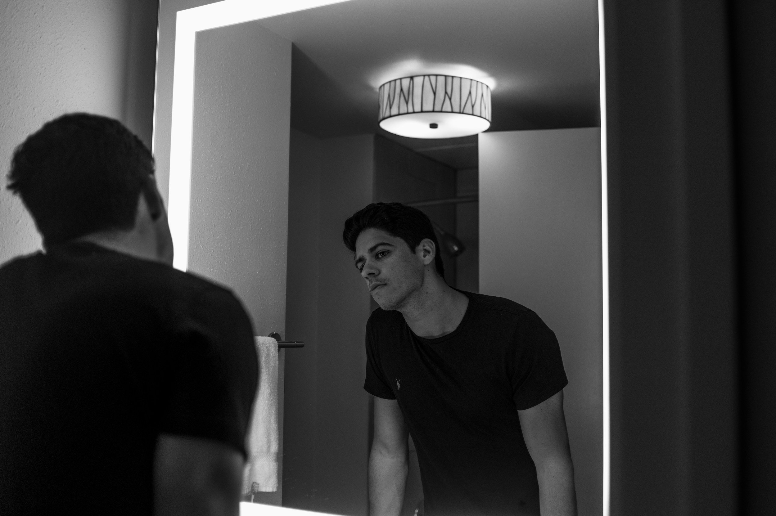 A young man looking at his reflection in a bathroom mirror, with a towel hanging on a towel rack behind him, illuminated by ceiling light.
