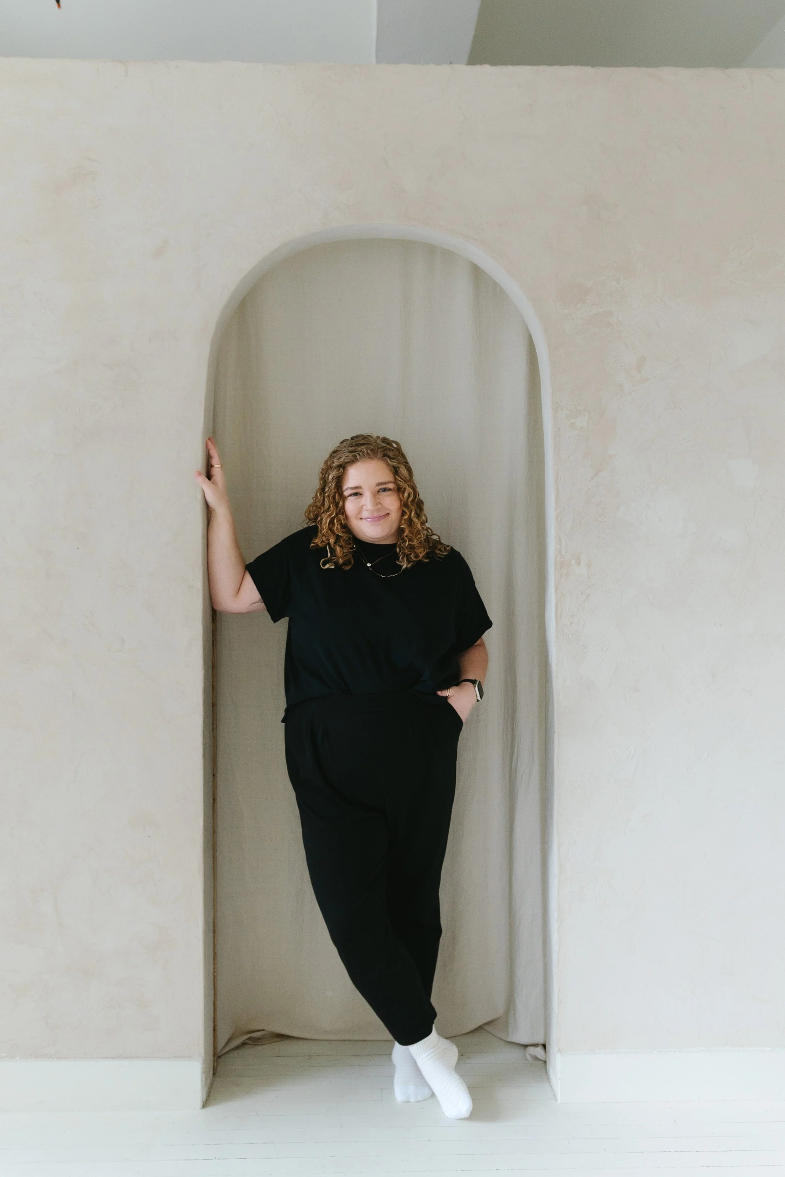 A woman with curly blonde hair standing in an arched doorway, smiling, wearing a black t-shirt, black pants, and white socks.