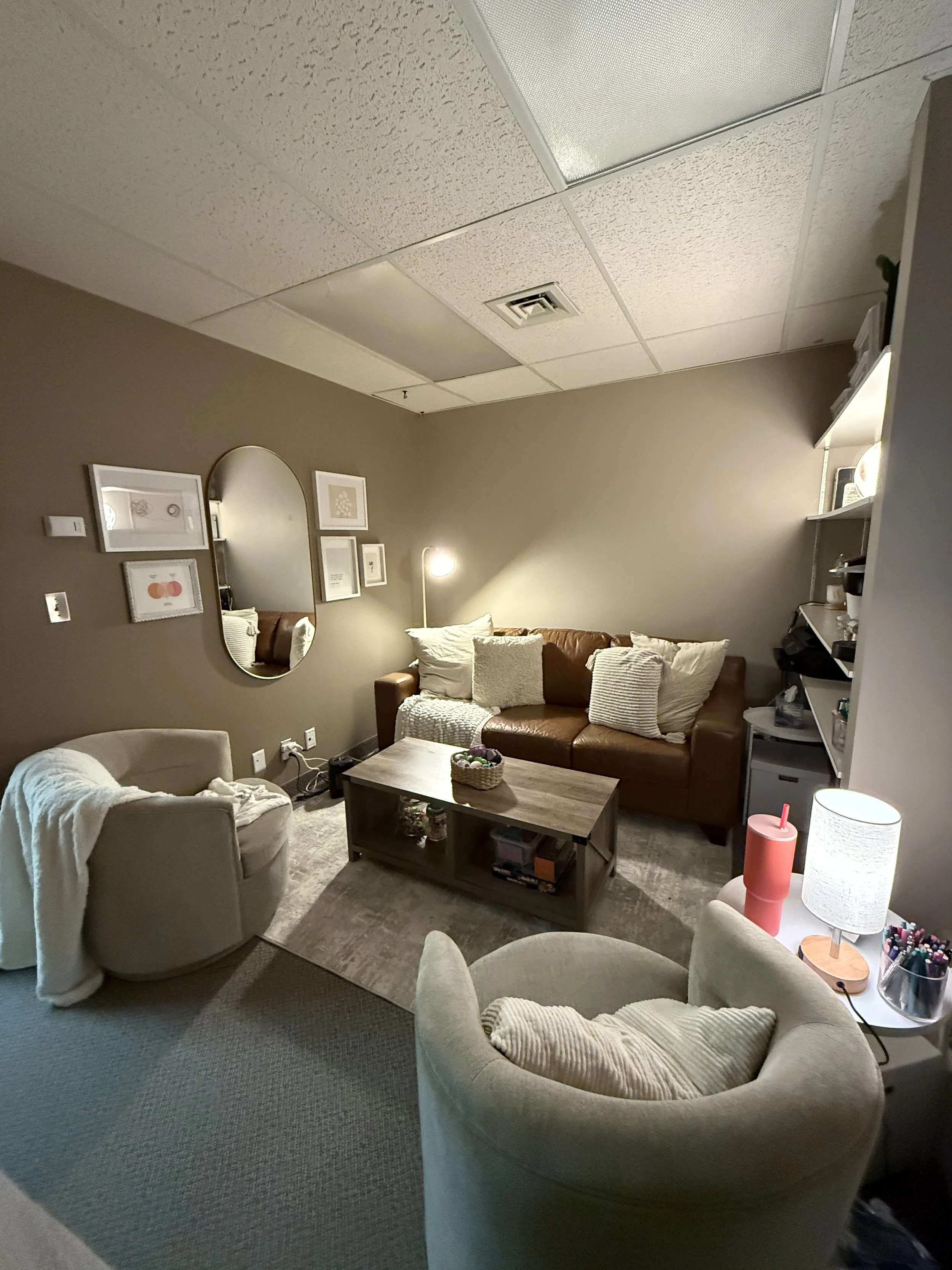 A cozy living room with a brown sofa decorated with white and striped pillows, a coffee table, a beige armchair with a blanket, a side table with a lamp, and wall art, with a corner shelf and a textured ceiling.