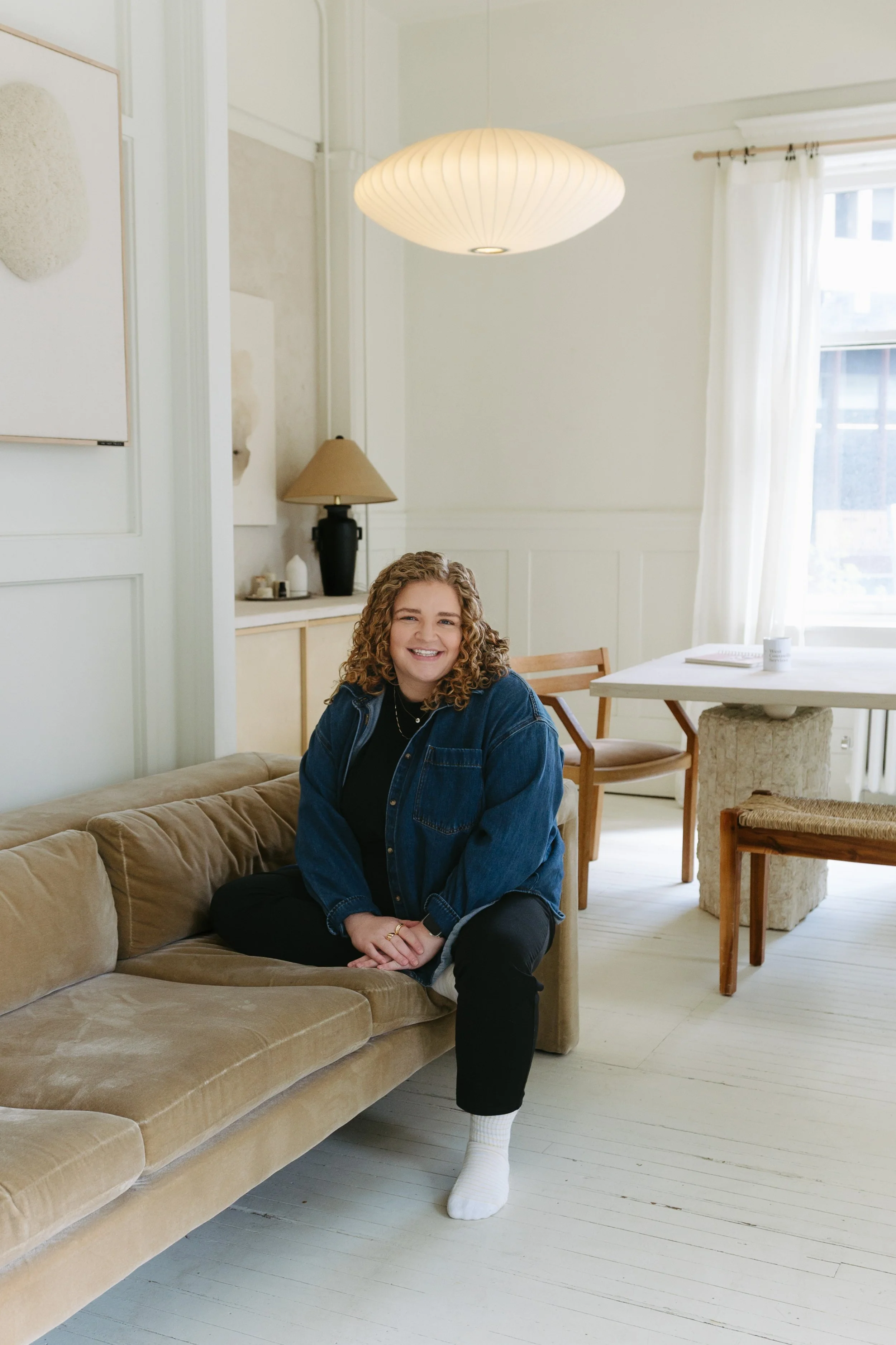 A young woman with curly hair, wearing a denim jacket, black pants, and white socks, sitting on a beige couch in a bright, minimalist room with white walls, art on the wall, a window with curtains, and wooden chairs and tables in the background.