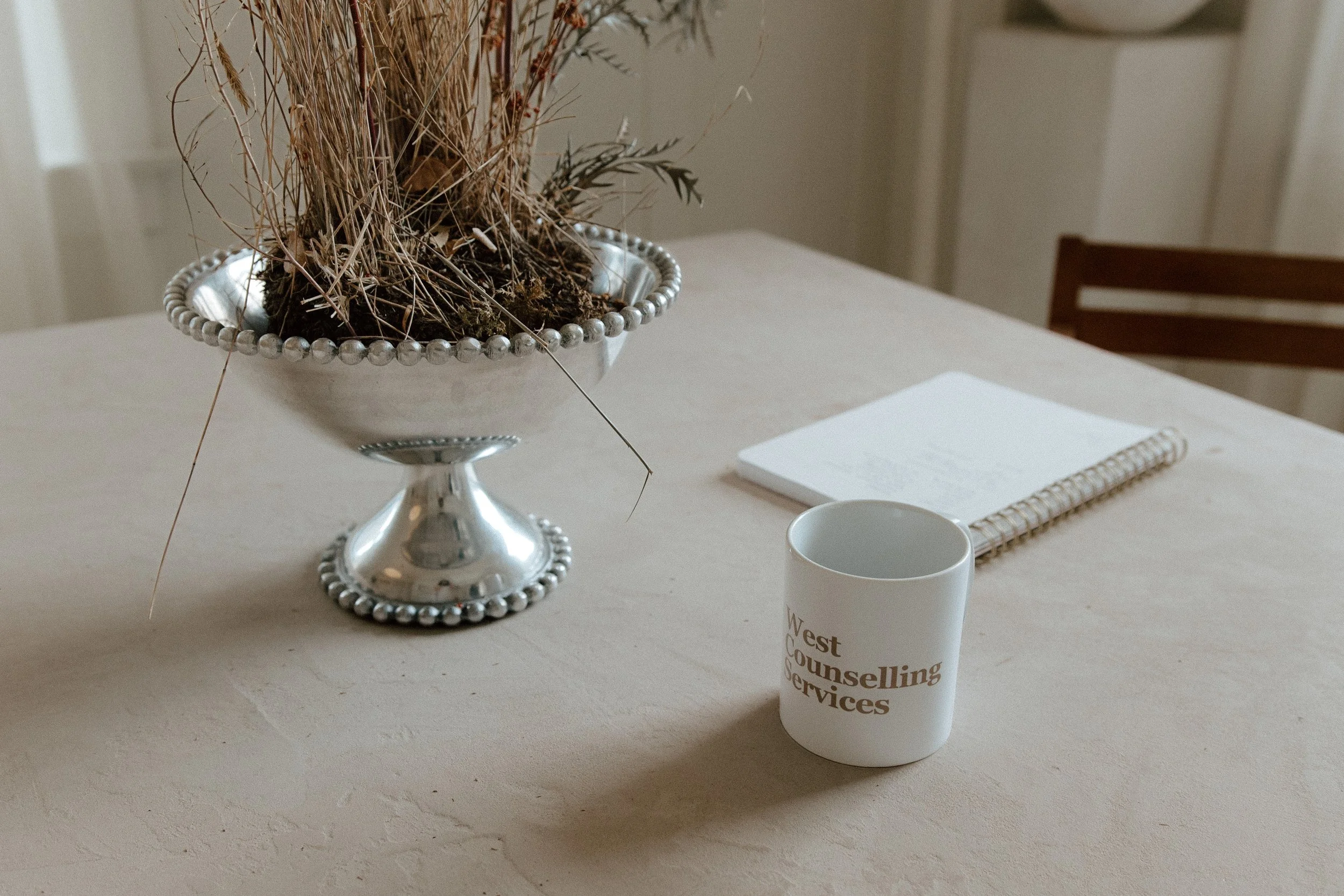 A table decorated with a silver bowl filled with dried flowers and grasses, a white coffee mug with text, a notebook, and a white napkin. The background shows a closed door and a wooden chair.