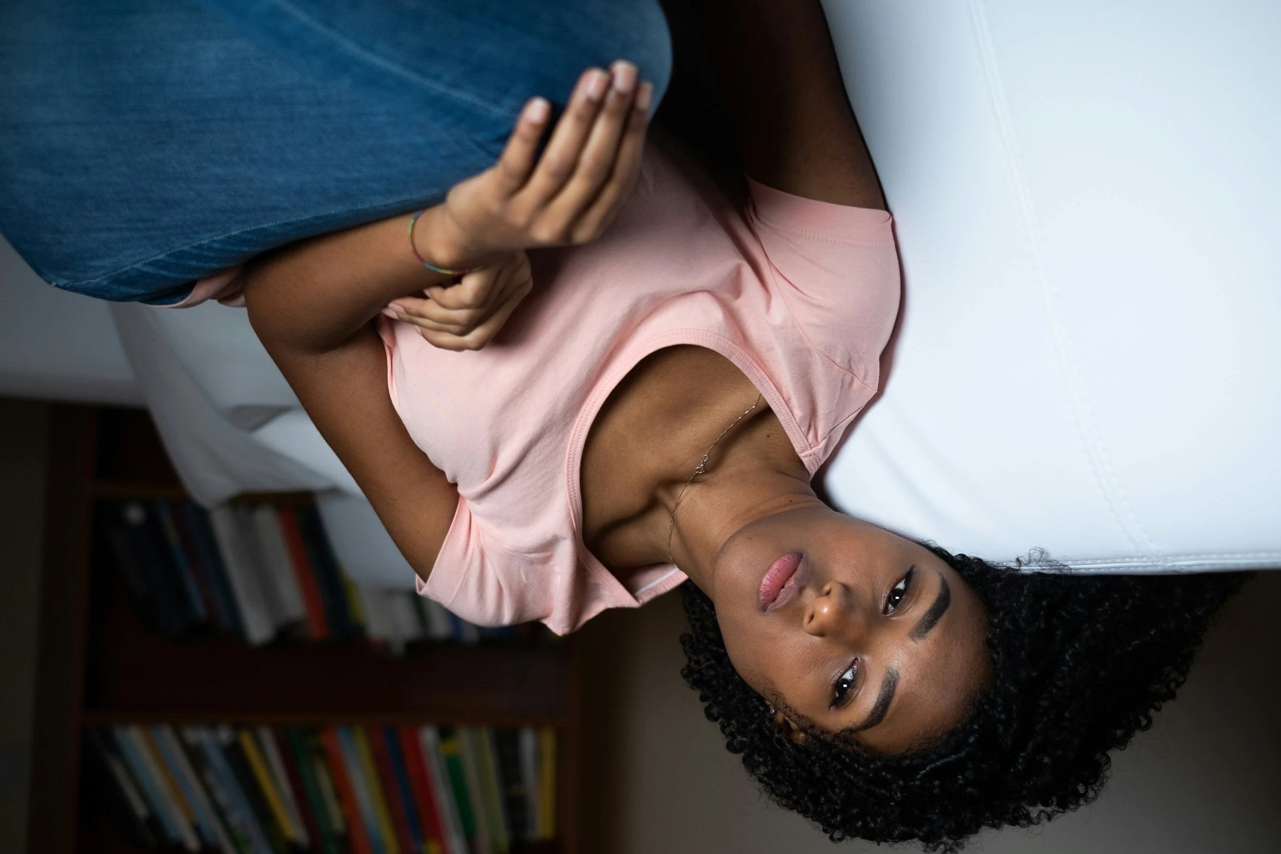 A woman with curly black hair and dark skin lying on a white couch, with her hand resting on her chest. She is wearing a peach sleeveless top and has a gentle expression on her face, with books on a shelf in the background.