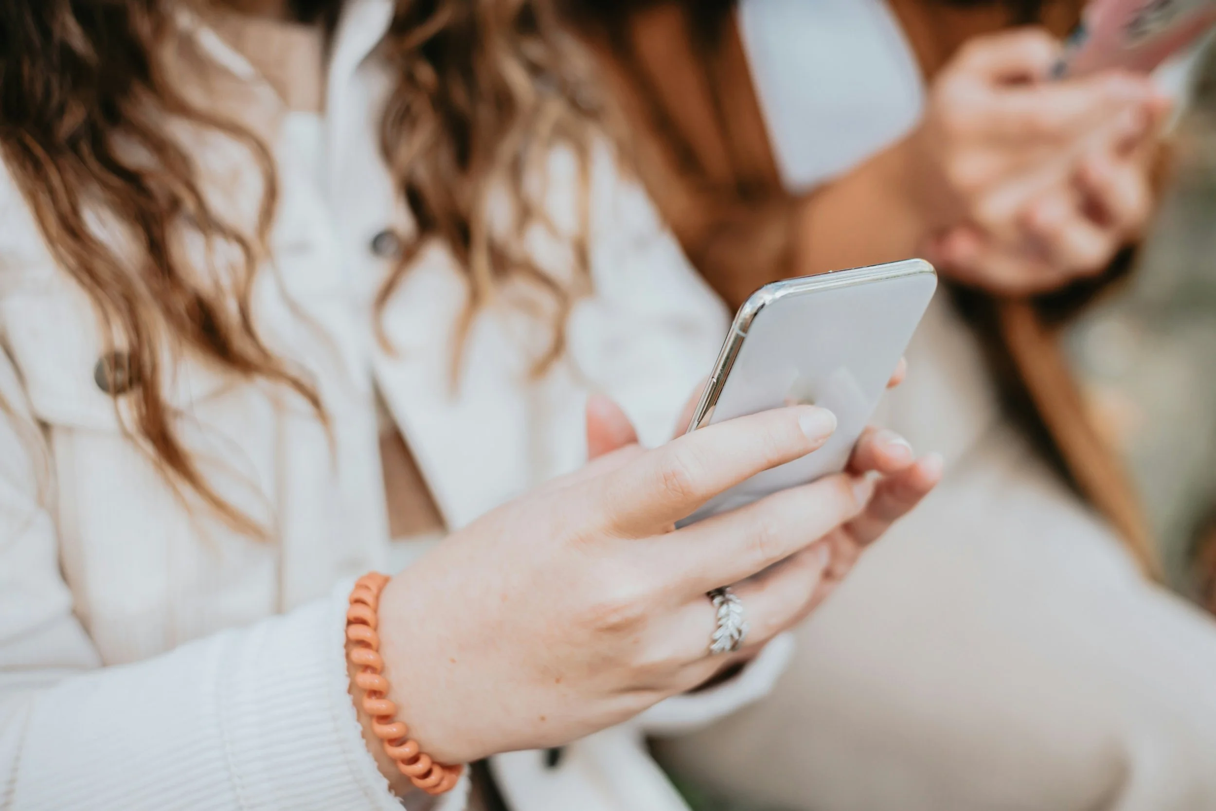 Close-up of a young woman with long, curly brown hair using her smartphone, with another person next to her also looking at a device, both wearing casual clothing and bracelets.