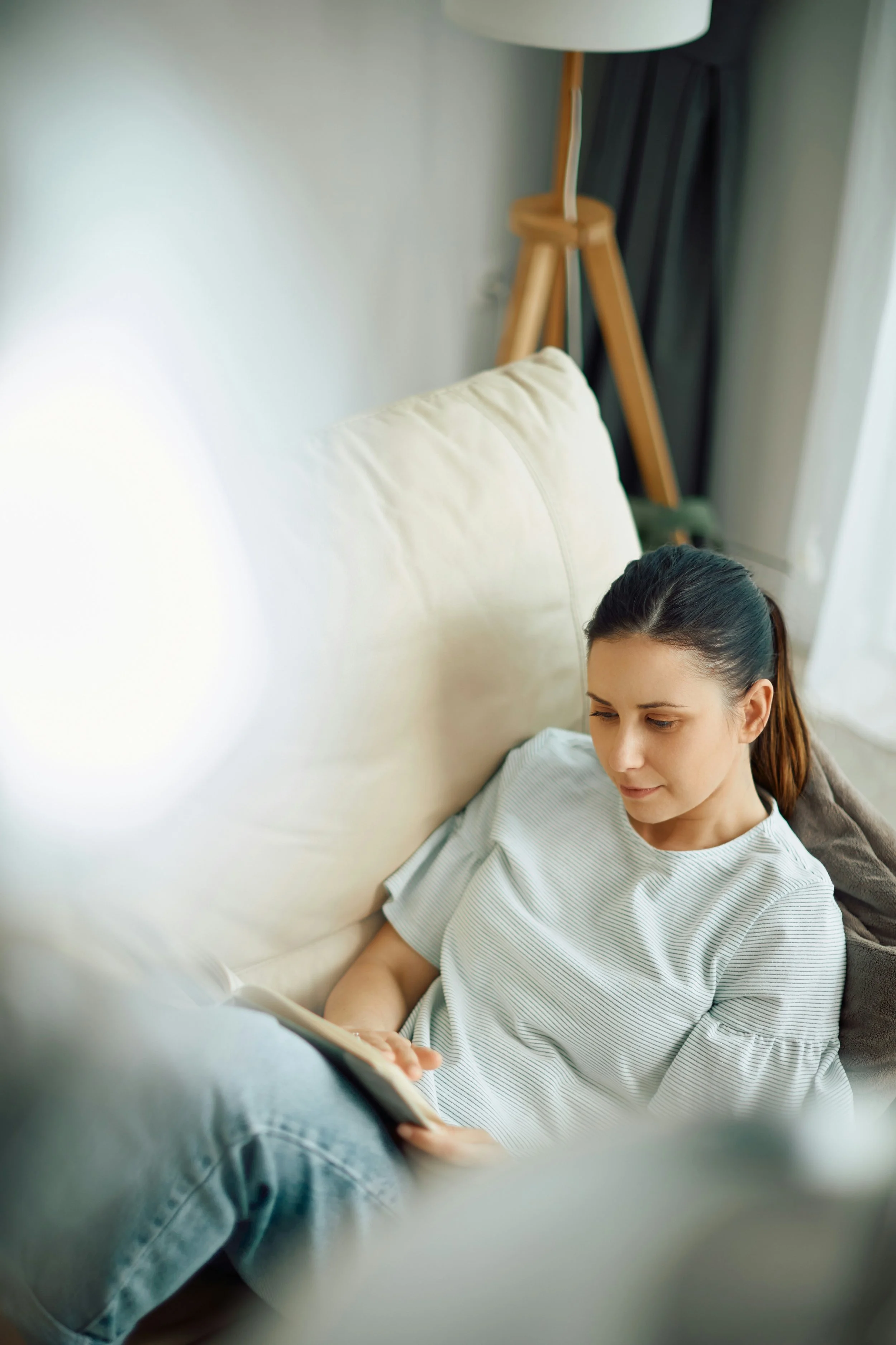 A young woman with long brown hair tied back, lying on a white couch, reading a book, wearing a striped white and gray t-shirt, with a cream pillow behind her and a wooden tripod lamp in the background.