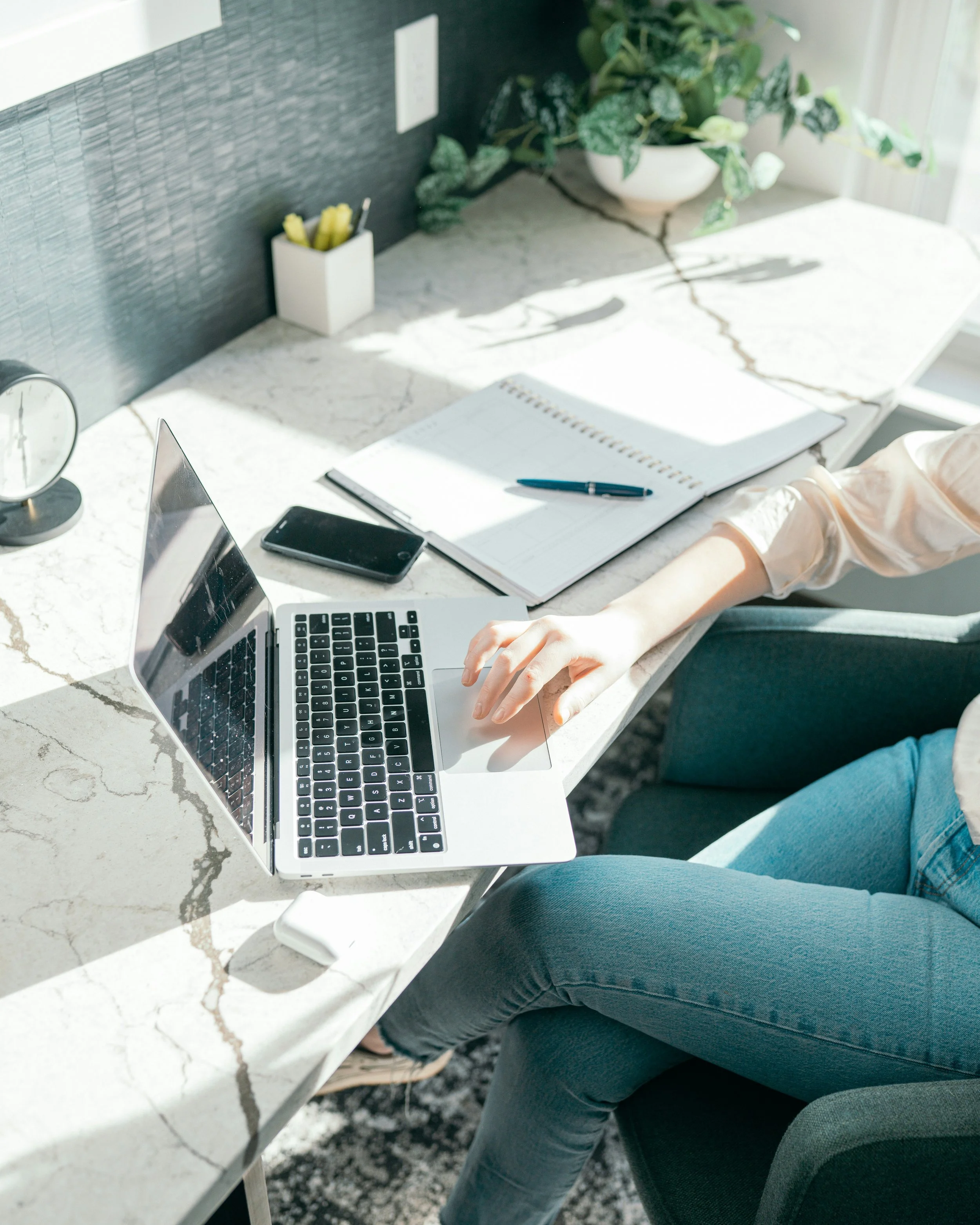 Person working at a white marble desk with a laptop, smartphone, notebook, a pen, a clock, and a container of pens. There is a potted plant and some electrical outlets in the background.