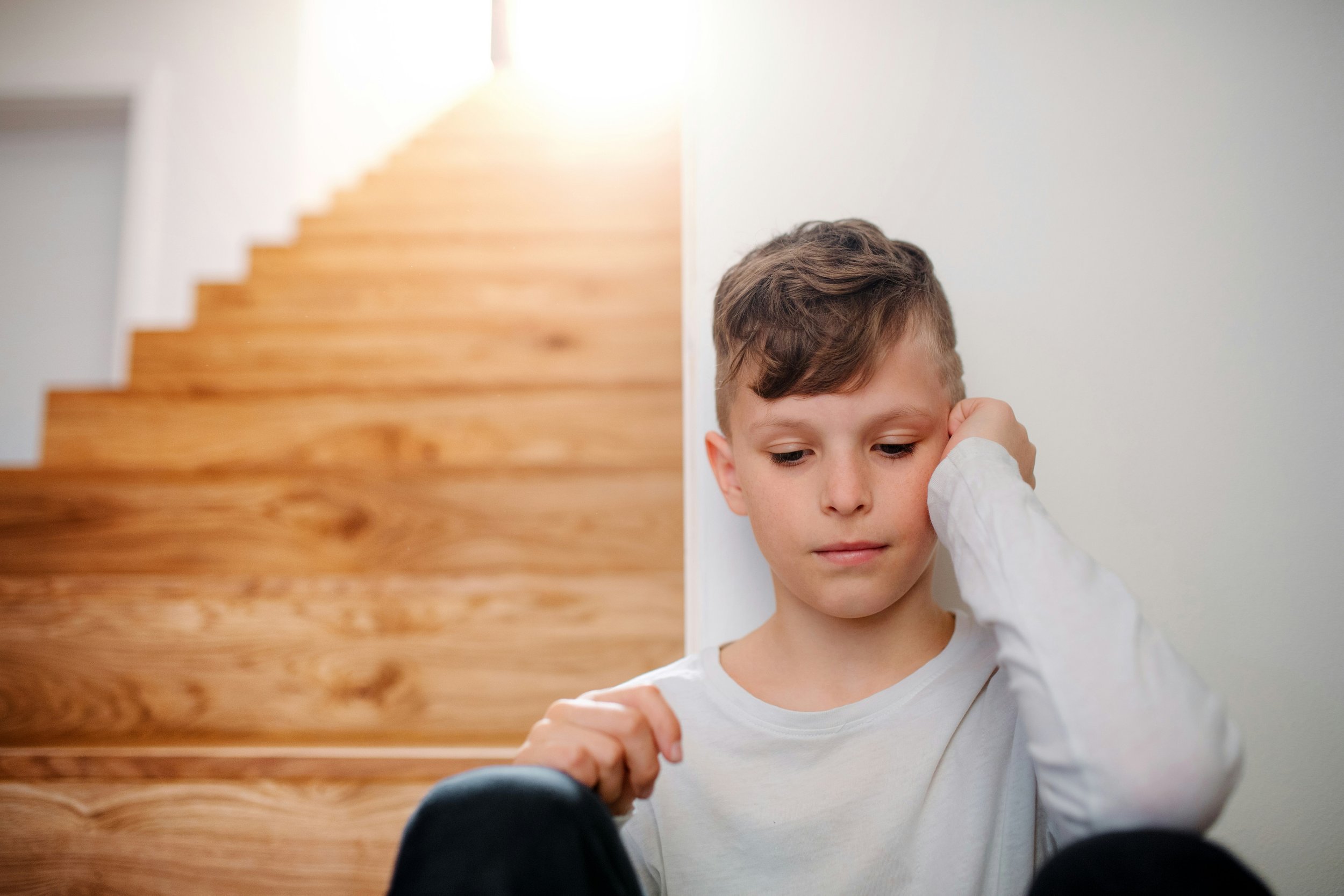 A young boy with brown hair, wearing a white shirt, standing near a white wall, resting his head on his hand, with a staircase with wooden steps in the background.