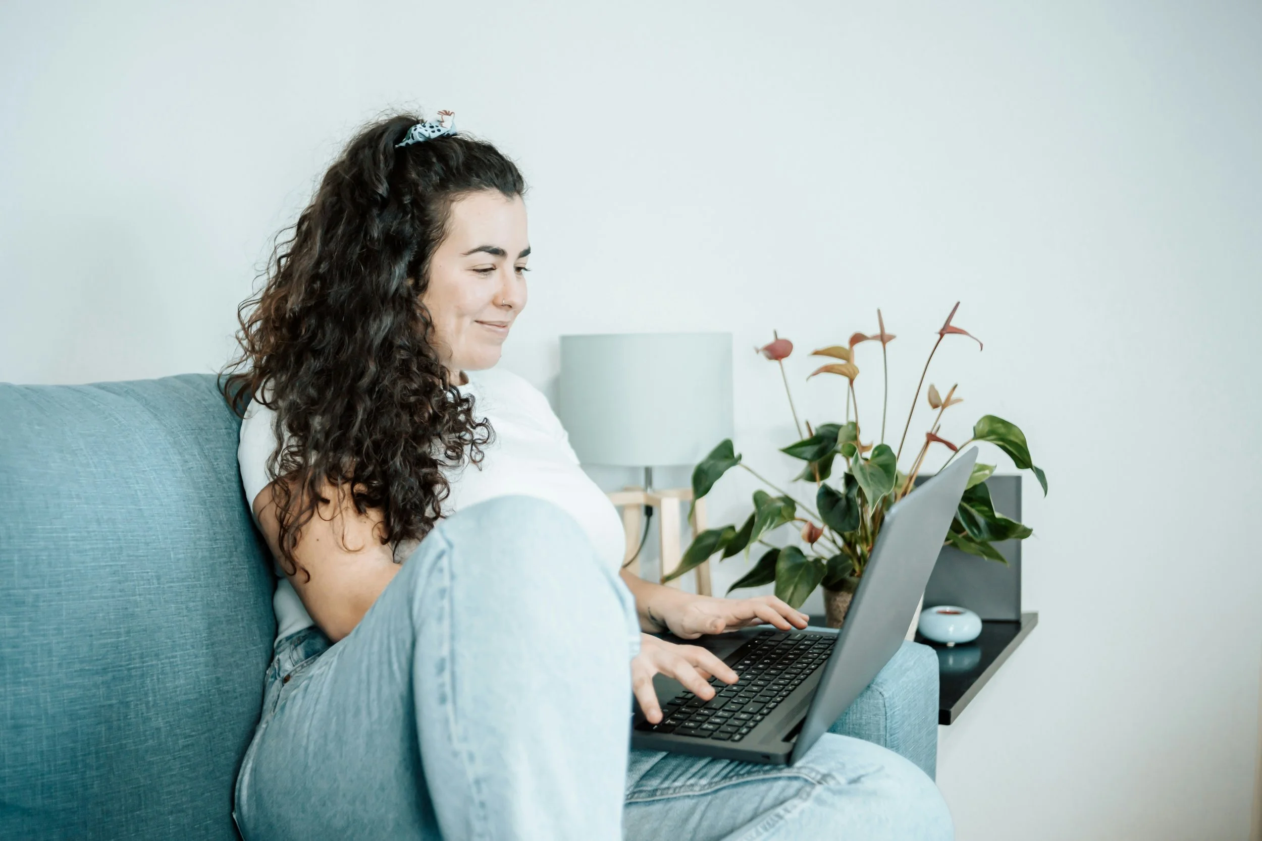 A young woman with long curly hair, wearing a white top and light blue jeans, sits on a blue couch using a laptop. There is a potted plant with green leaves and some pinkish flowers nearby, a white lamp, and a small white object on a side table in a bright, minimalist room.
