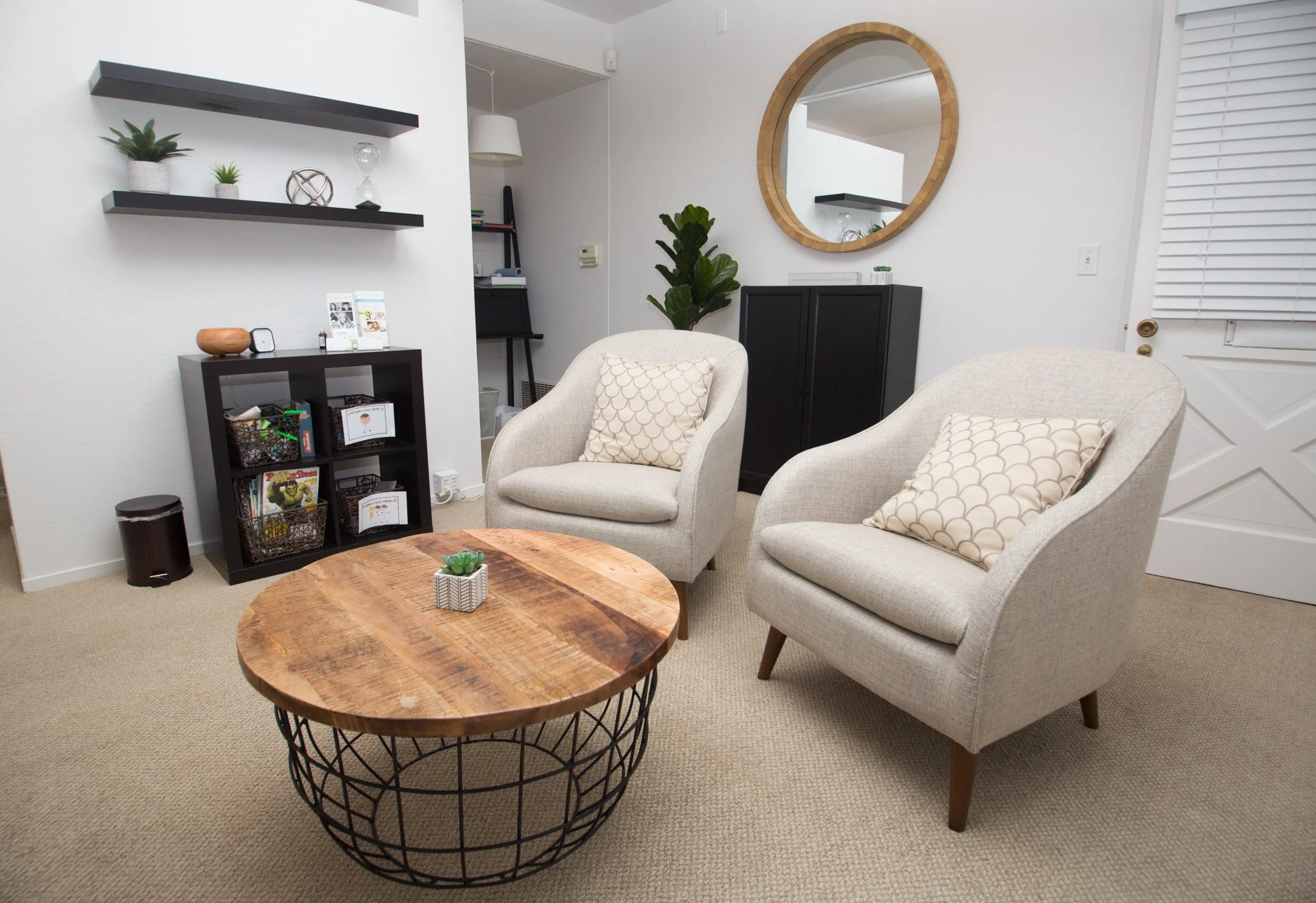 Living room with two beige armchairs with patterned pillows, a wooden coffee table with a small plant, black shelves with decorative items, a large green potted plant, a wall mirror, and various small furnishings.