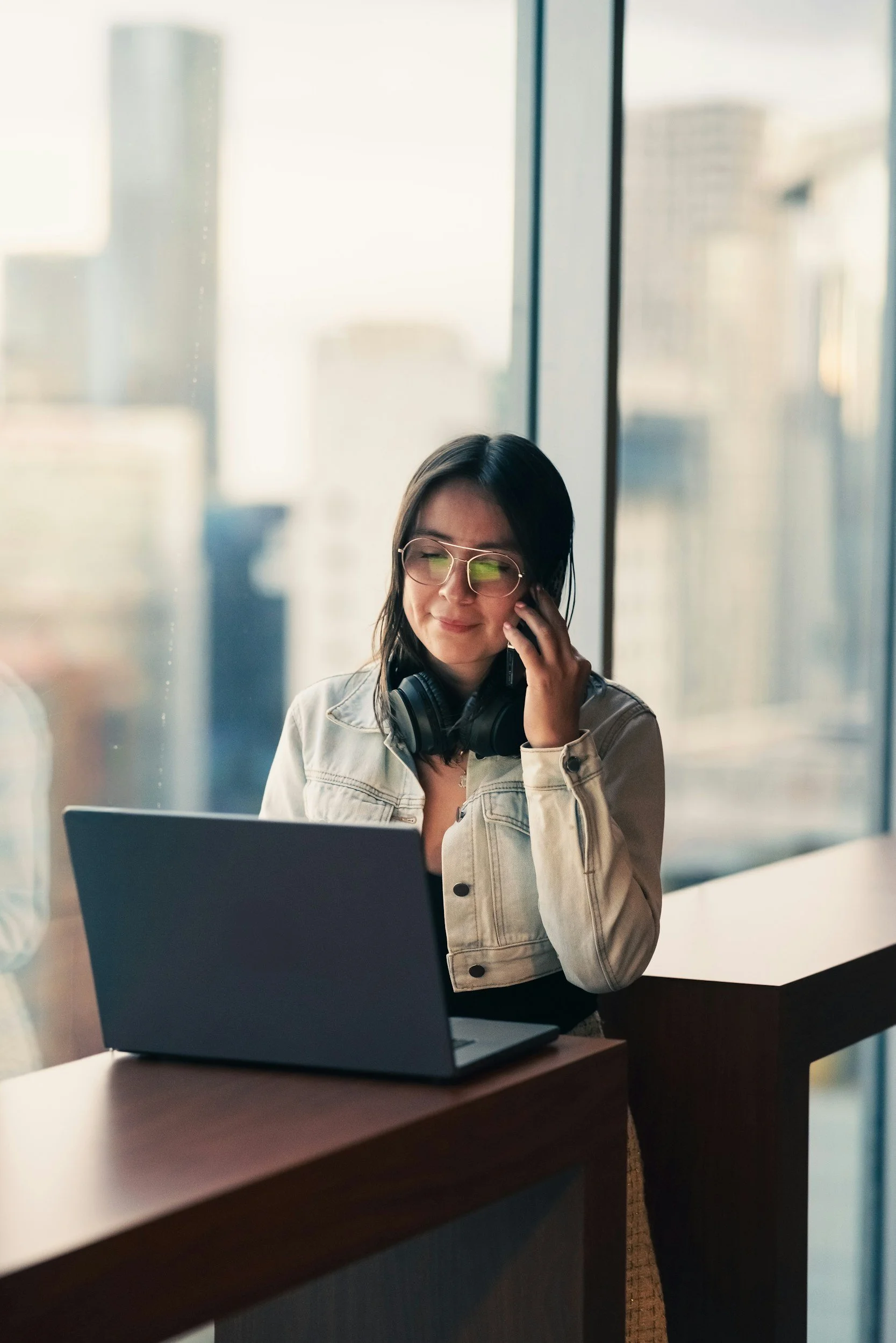 A woman with glasses and headphones around her neck sitting at a desk, talking on the phone, with a laptop in front of her and large windows behind her showing a cityscape.