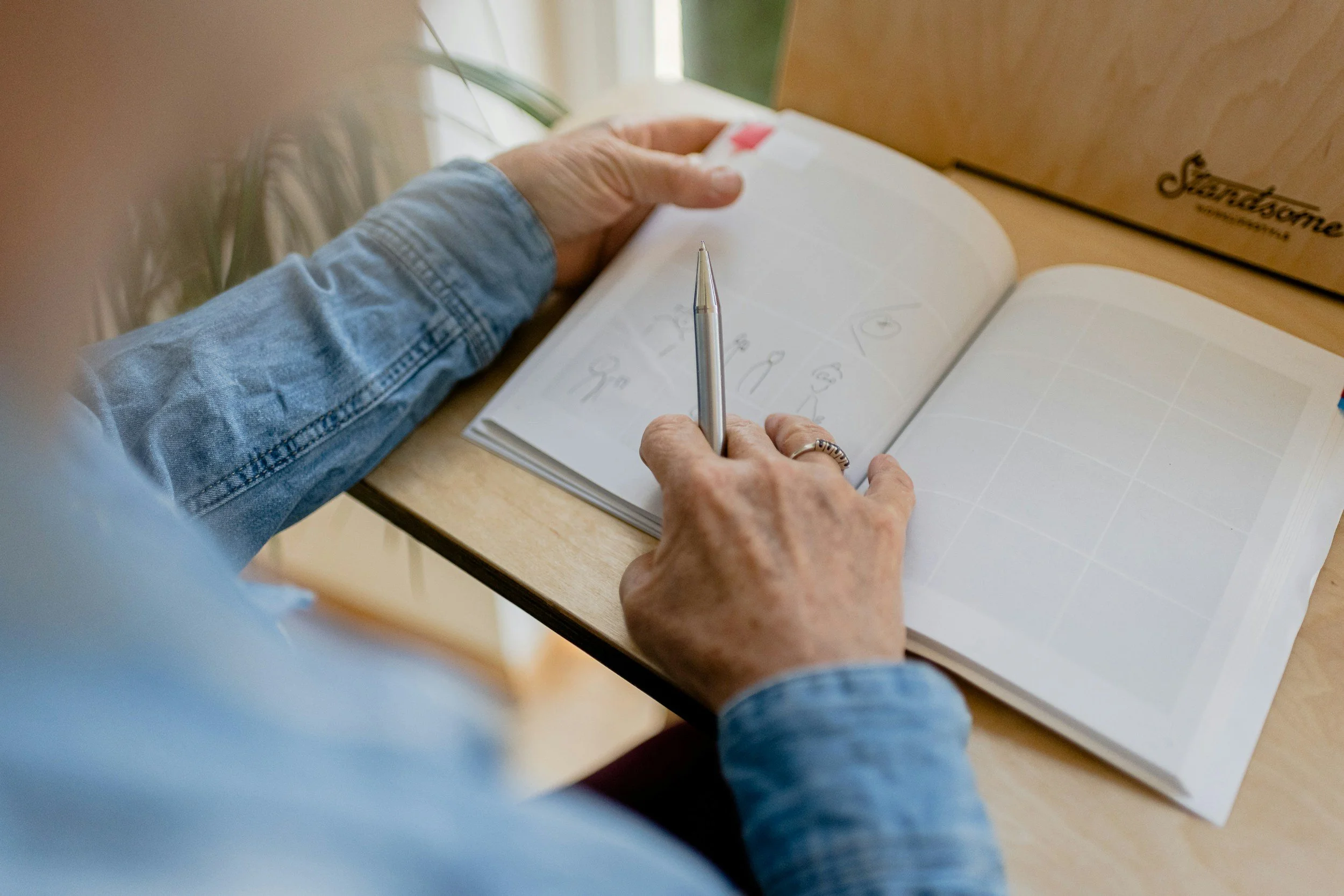 Person wearing a denim jacket writing in a notebook with a pen on a wooden table, with a cardboard box labeled 'Saxony' nearby.