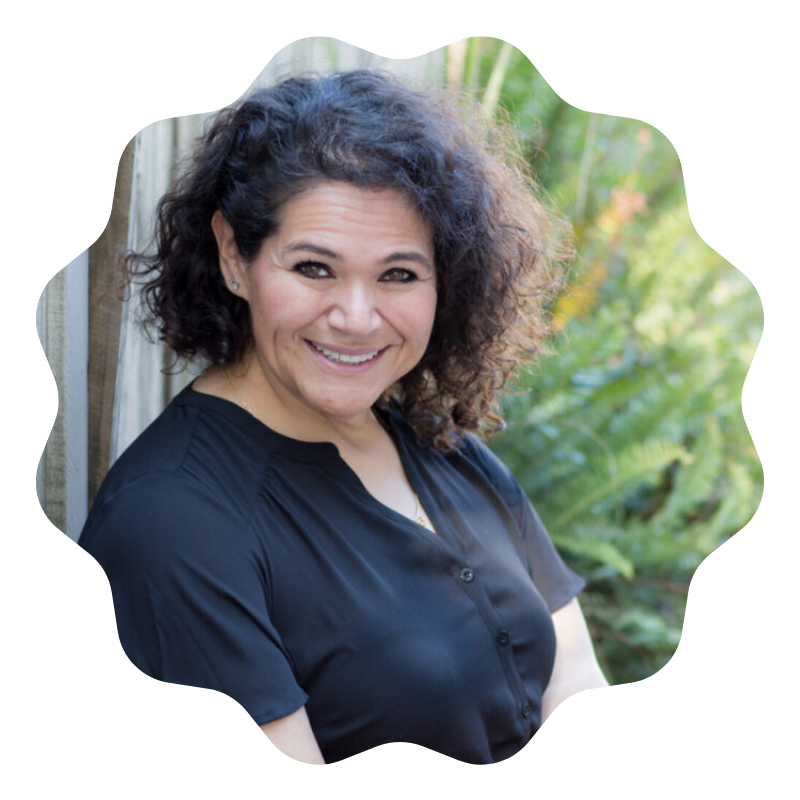 A woman with curly dark hair smiling outdoors in front of green plants, wearing a black shirt.