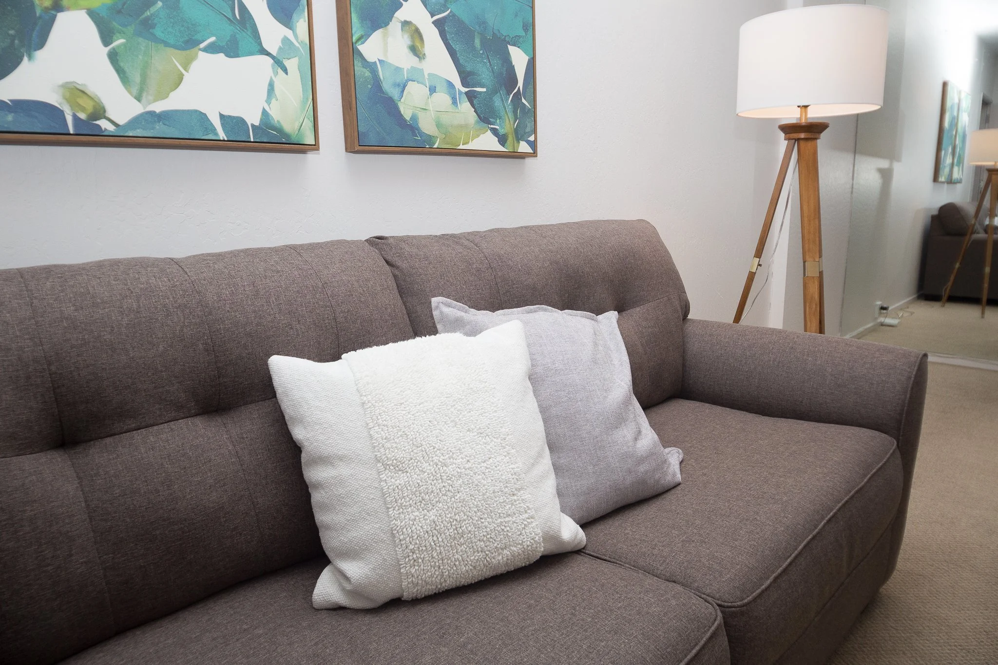 Living room with a brown fabric sofa decorated with a white textured pillow and a small beige pillow, above the sofa are two framed botanical art prints featuring green leaves. To the right of the sofa is a wooden tripod floor lamp with a white lampshade, and in the background is a full-length mirror reflecting part of the room and a hint of furniture.