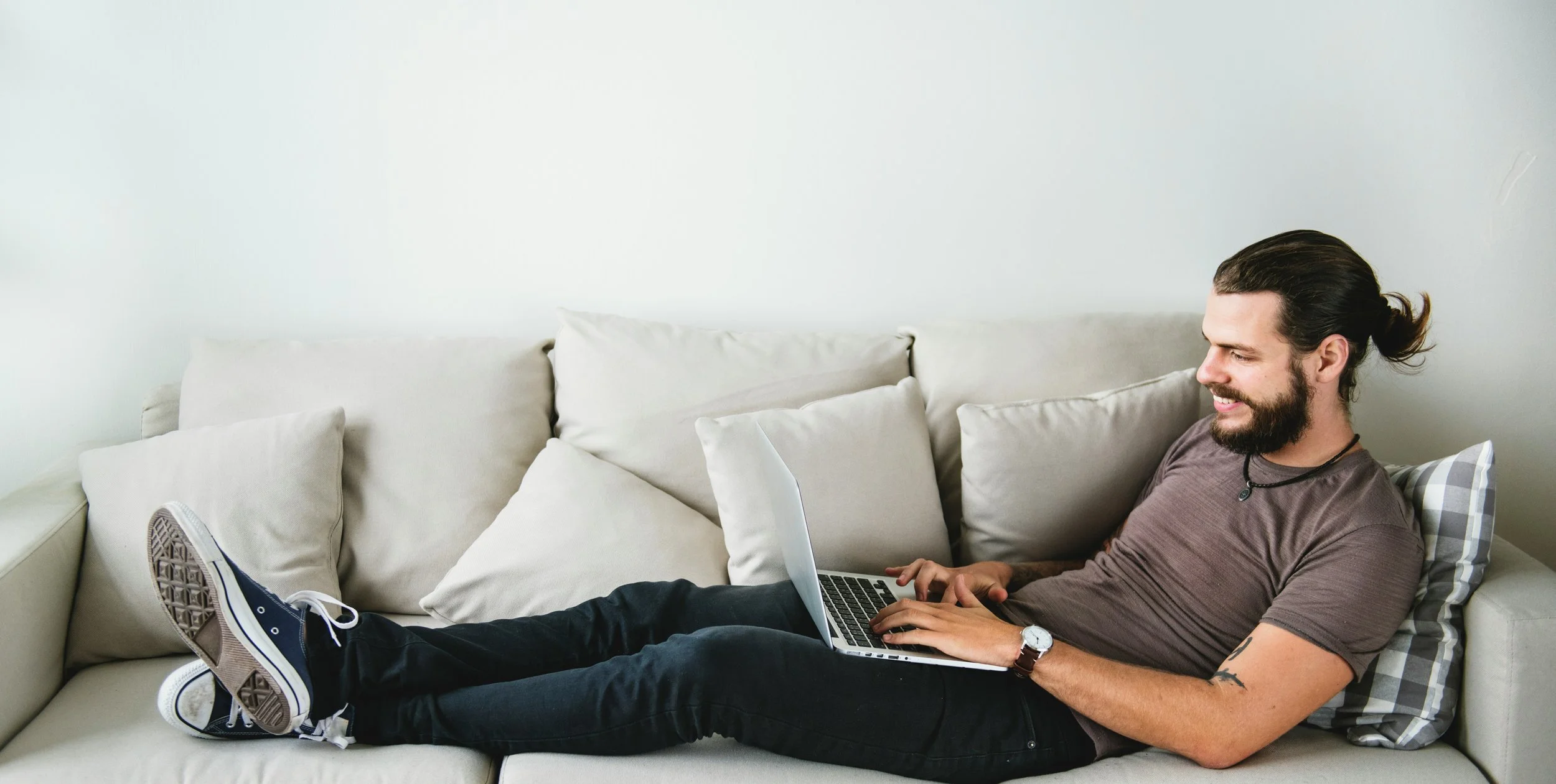 A man with long hair and a beard sitting on a beige sofa, smiling and using a laptop, with multiple pillows around him.