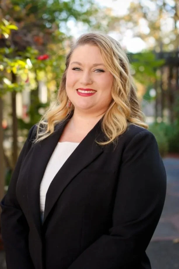 A woman with blonde wavy hair smiling, wearing a black blazer over a white top, outdoors with trees and greenery in the background.