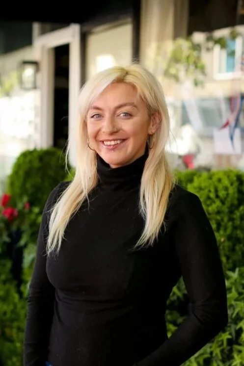 A smiling blonde woman in a black turtleneck standing outdoors with greenery and a building in the background.