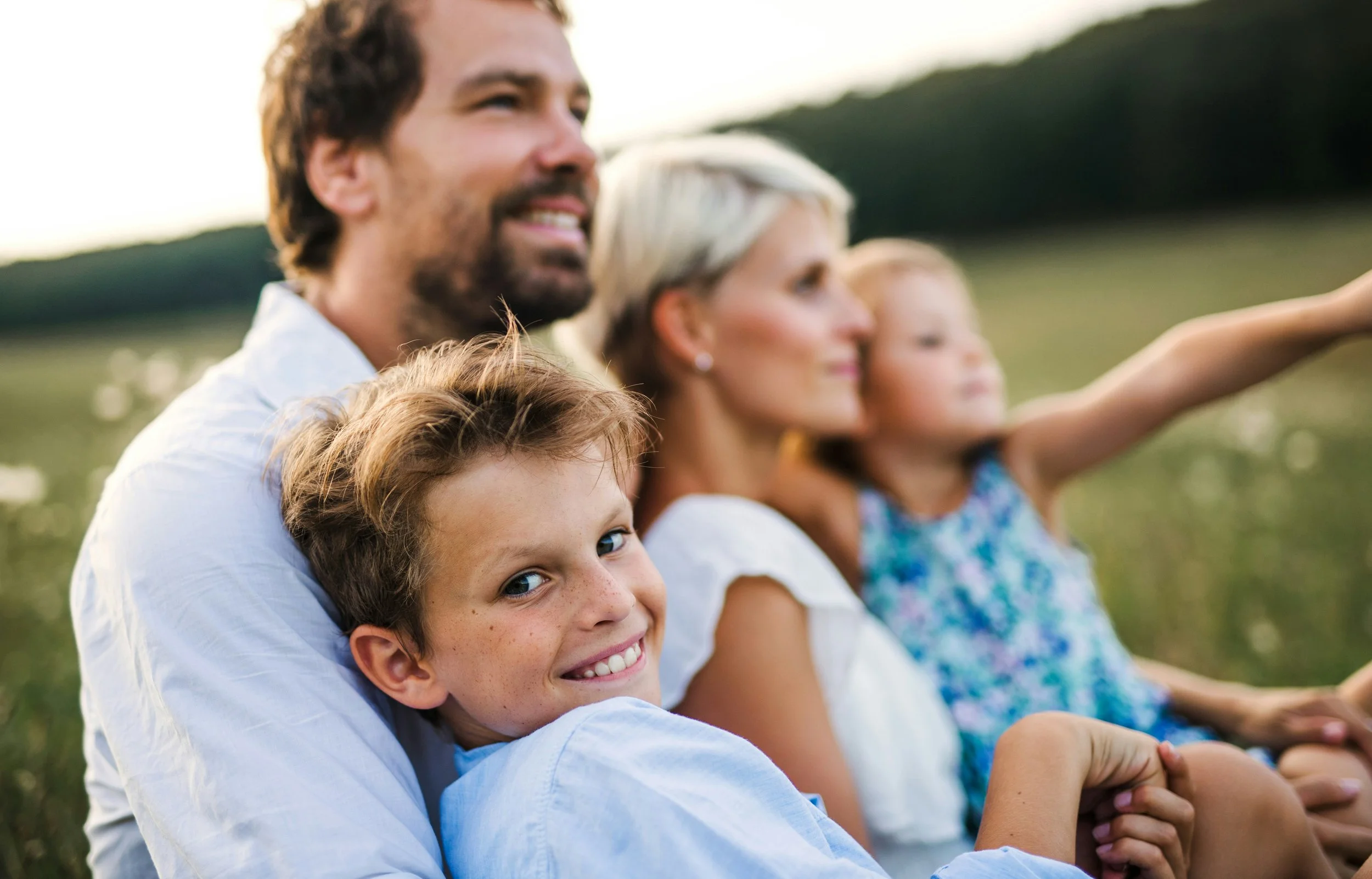 A happy family of four, including a man, two women, and a young girl, sitting outdoors in a field at sunset, smiling and enjoying the moment.