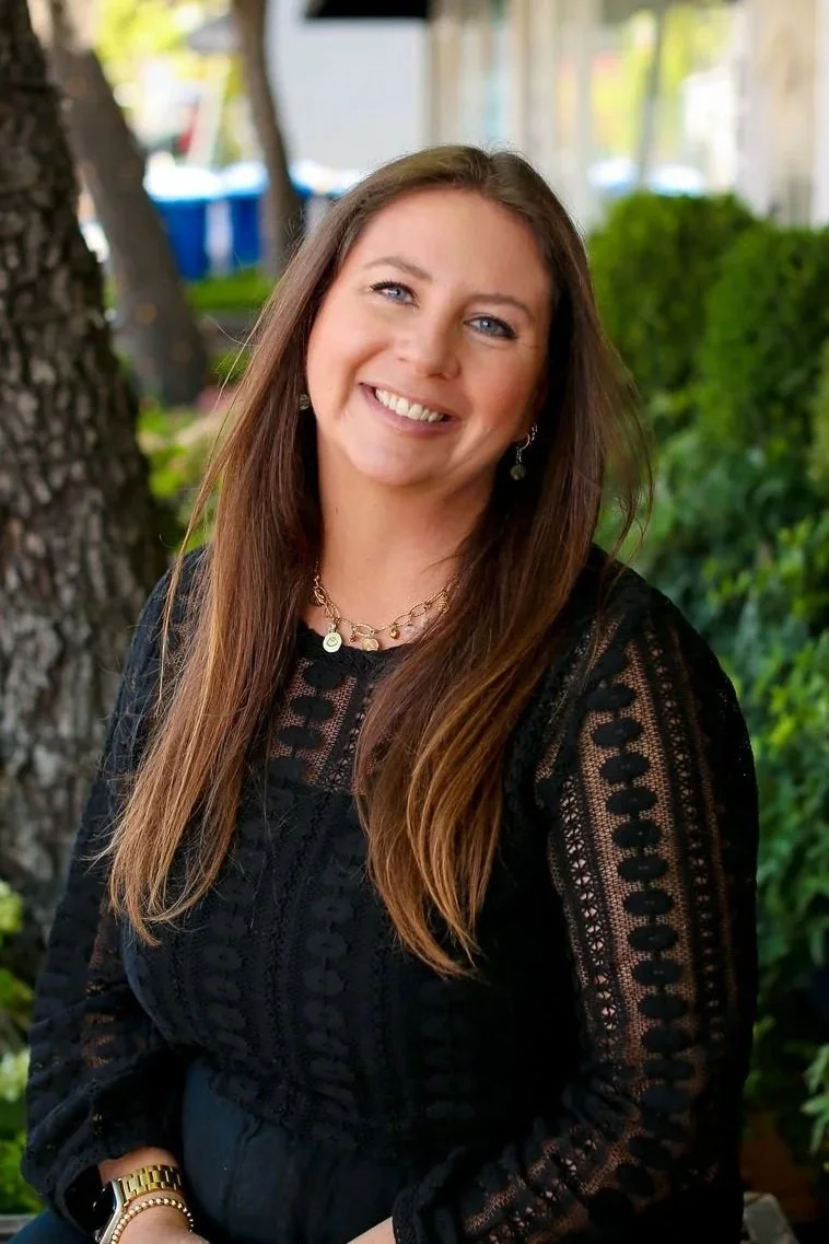 A woman with long brown hair and blue eyes smiling outdoors, wearing a black lace top, a gold watch, a necklace, and earrings, with trees and greenery in the background.