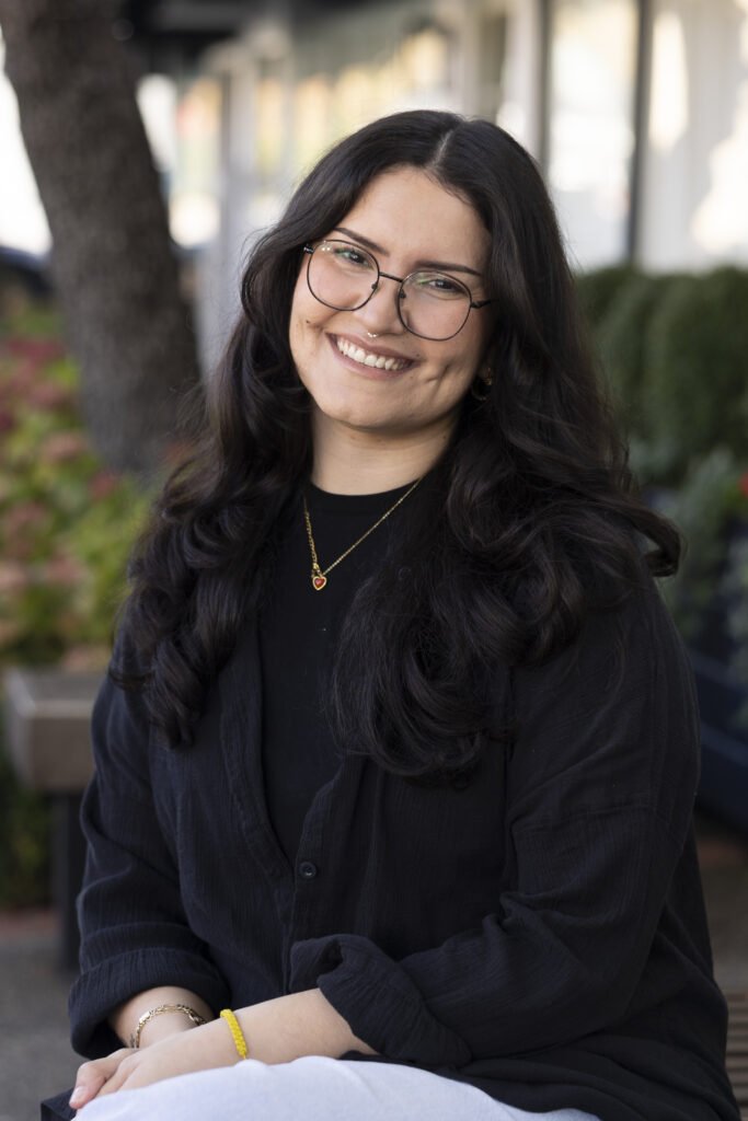 Young woman with long dark wavy hair, glasses, and a nose piercing, smiling outdoors in front of trees and plants, wearing a black shirt and jewelry.