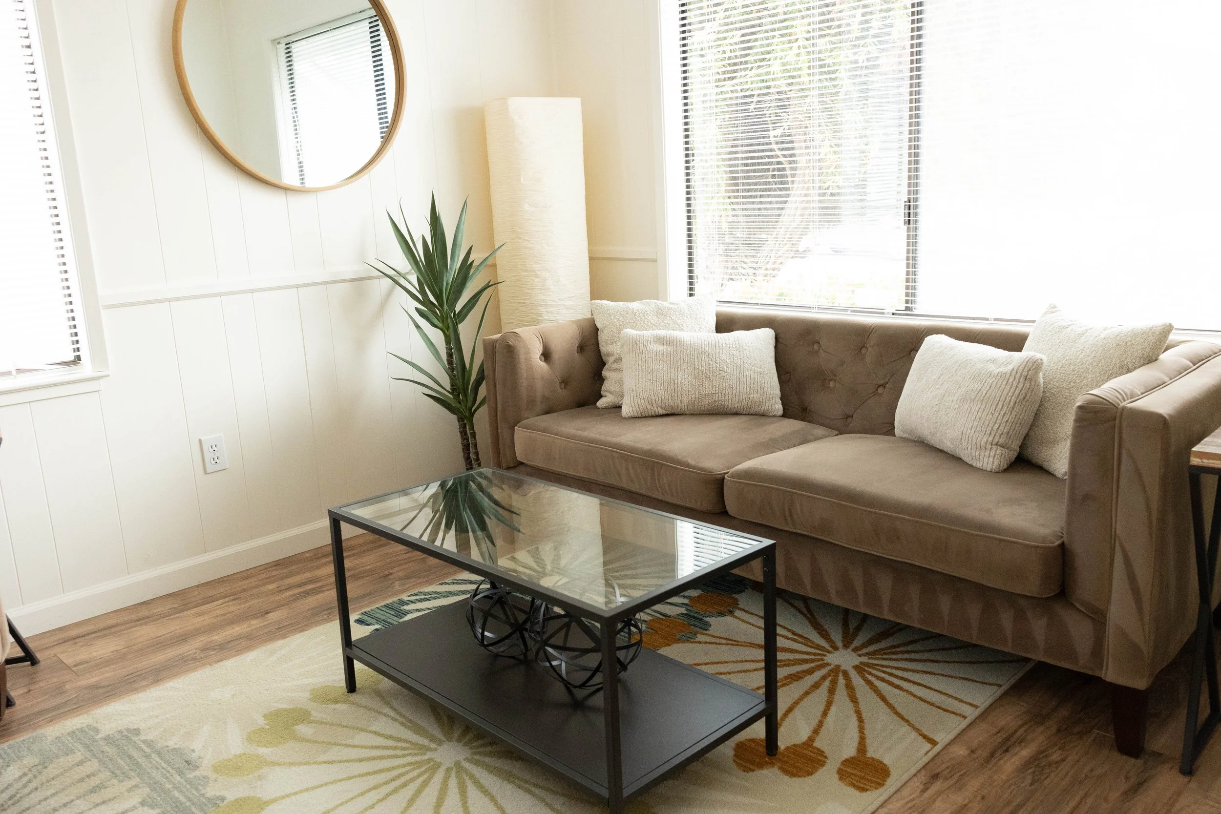 Living room with a beige tufted couch, white pillows, a tall white floor lamp, a green houseplant, a glass coffee table with decorative objects, a colorful area rug, and windows with blinds.