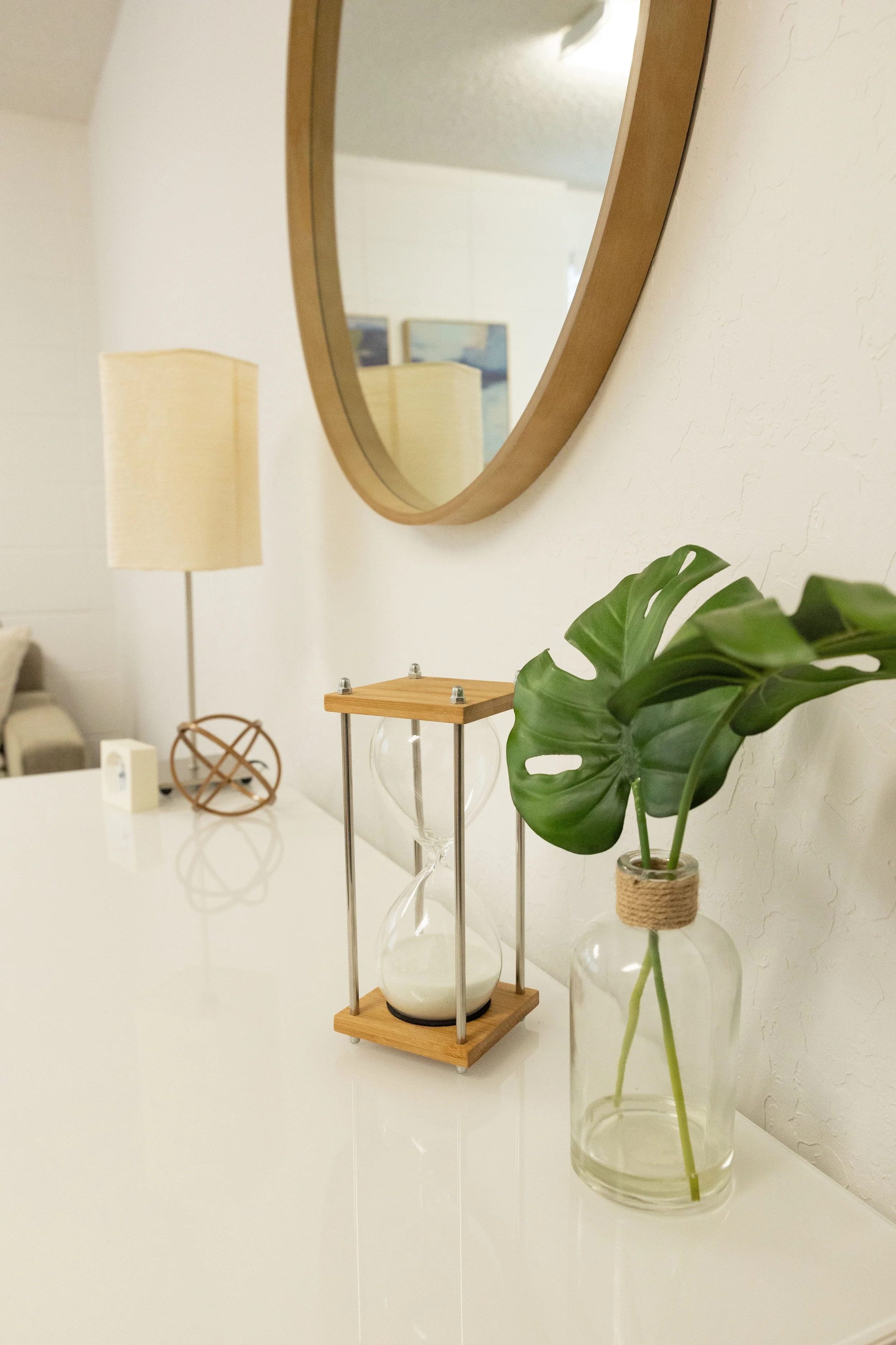Decorative display with a potted monstera plant in a glass vase wrapped with twine, a wooden hourglass, and a wooden shelf with a glass base on a white surface. In the background, a wall mirror and a standing lamp are visible.