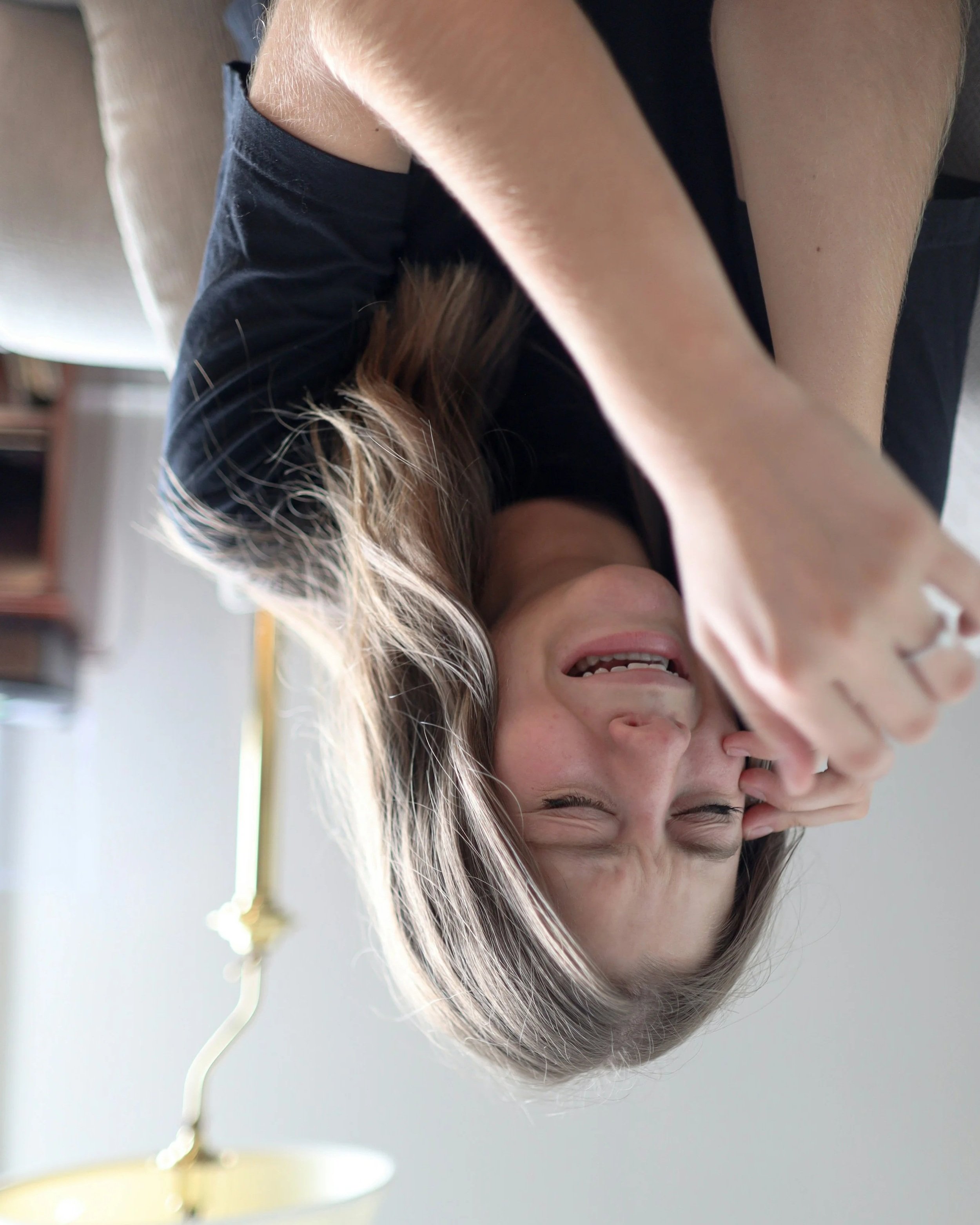Upside-down close-up of a woman laughing with her eyes closed, showing her teeth, with long hair and a hand near her face, background with a lamp and blurred objects.