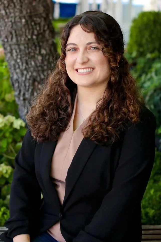 A woman with curly brown hair smiling outdoors with greenery and a tree in the background.