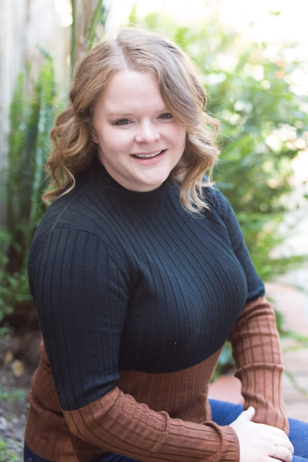 A woman with curly blonde hair smiling outdoors in a garden setting, wearing a black and brown ribbed sweater.
