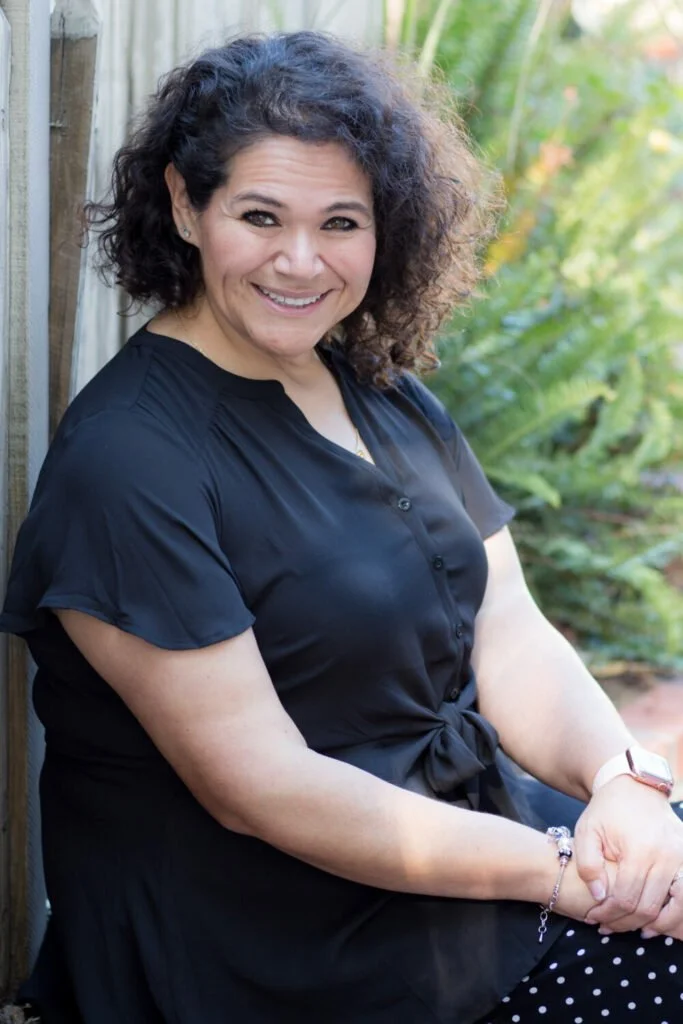 Smiling woman with curly hair, wearing a black dress, sitting outdoors near a wooden fence with plants in the background.