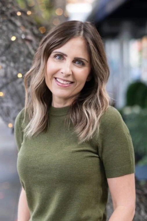 Headshot of a smiling woman with shoulder-length wavy brown hair, wearing a green shirt, standing outdoors with blurred background including lights and greenery.