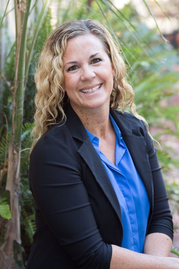 A woman with curly blonde hair smiling outdoors, wearing a black blazer and blue shirt, surrounded by green plants.