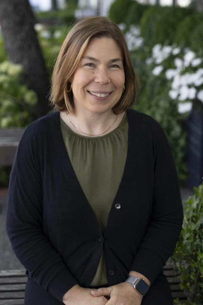 A woman with shoulder-length light brown hair, smiling, wearing a green top, black cardigan, gold hoop earrings, a silver watch, and a simple necklace, outdoors with lush green foliage in the background.