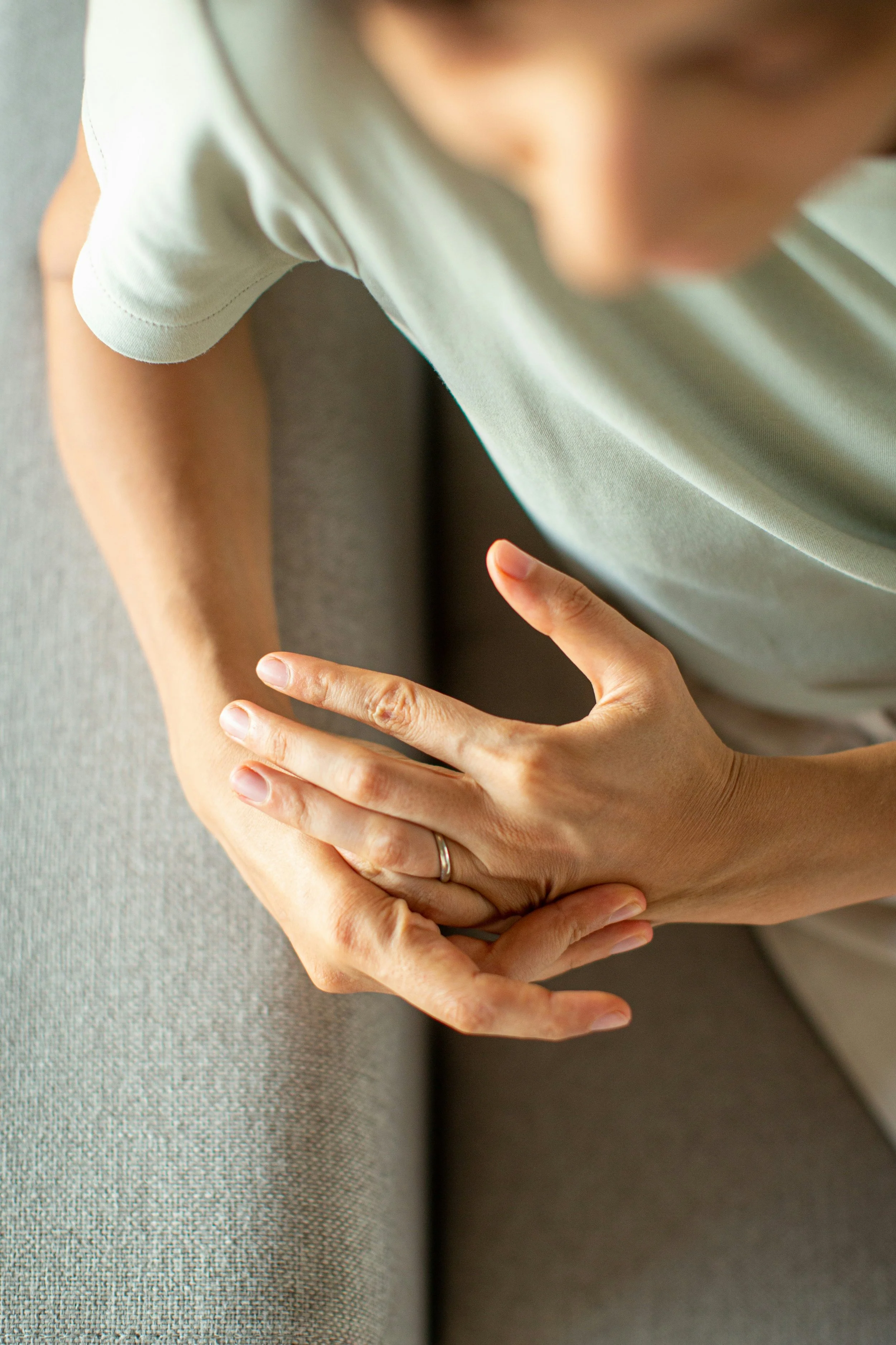 Close-up of a person's hand resting on their arm, wearing a ring, with a second hand pointing at it, on a gray fabric surface.