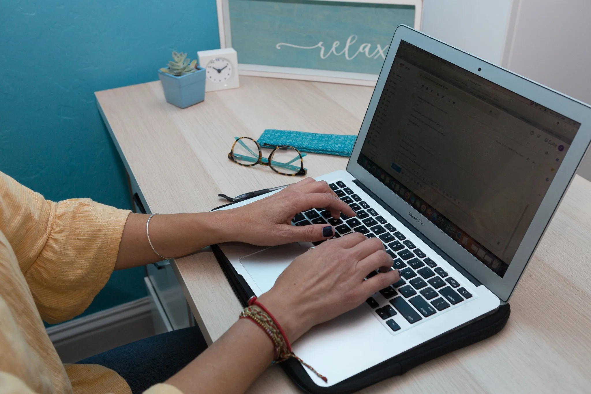 Person typing on a MacBook Air at a desk with glasses, a potted plant, a clock, and a framed sign that says 'relax'.