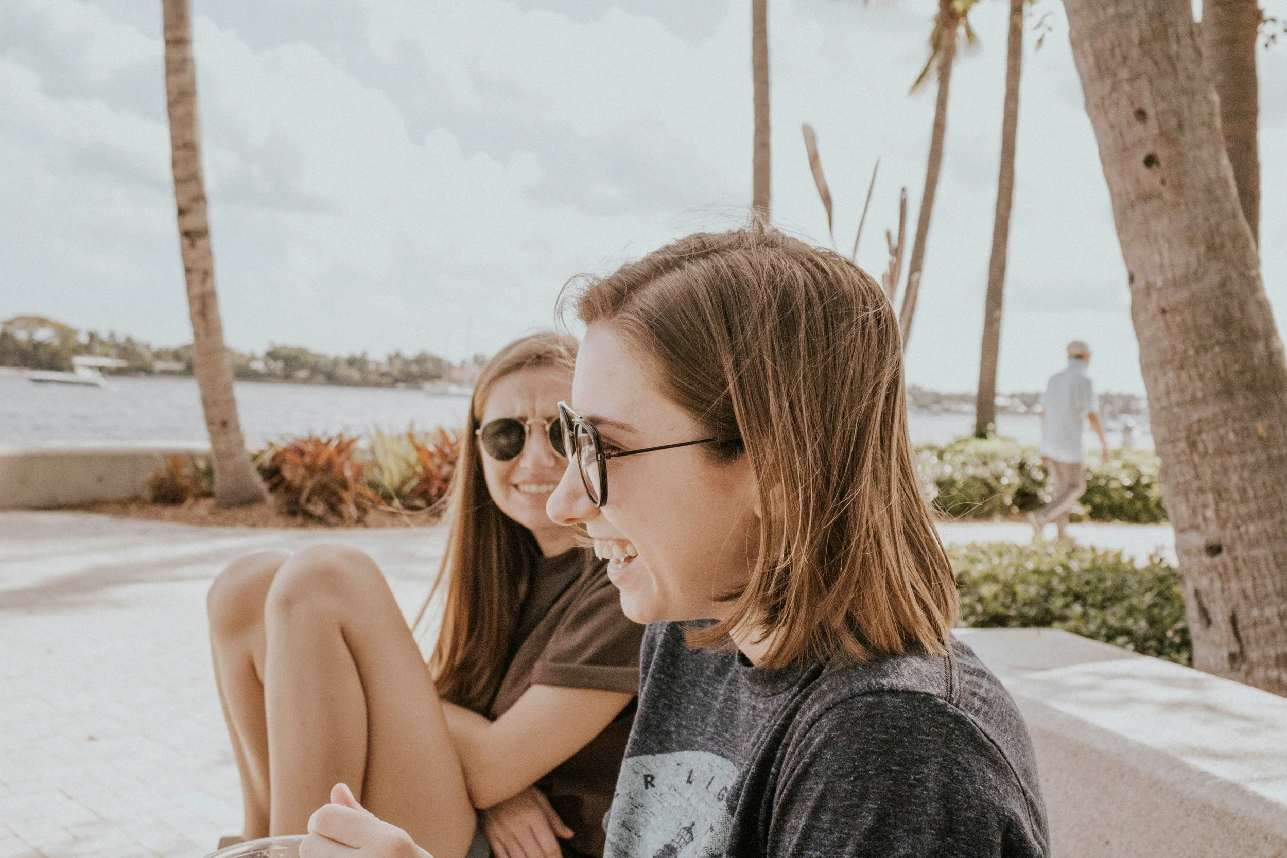 Two women with brown hair and glasses sitting outdoors near palm trees, smiling and laughing, with a body of water and a boat in the background.