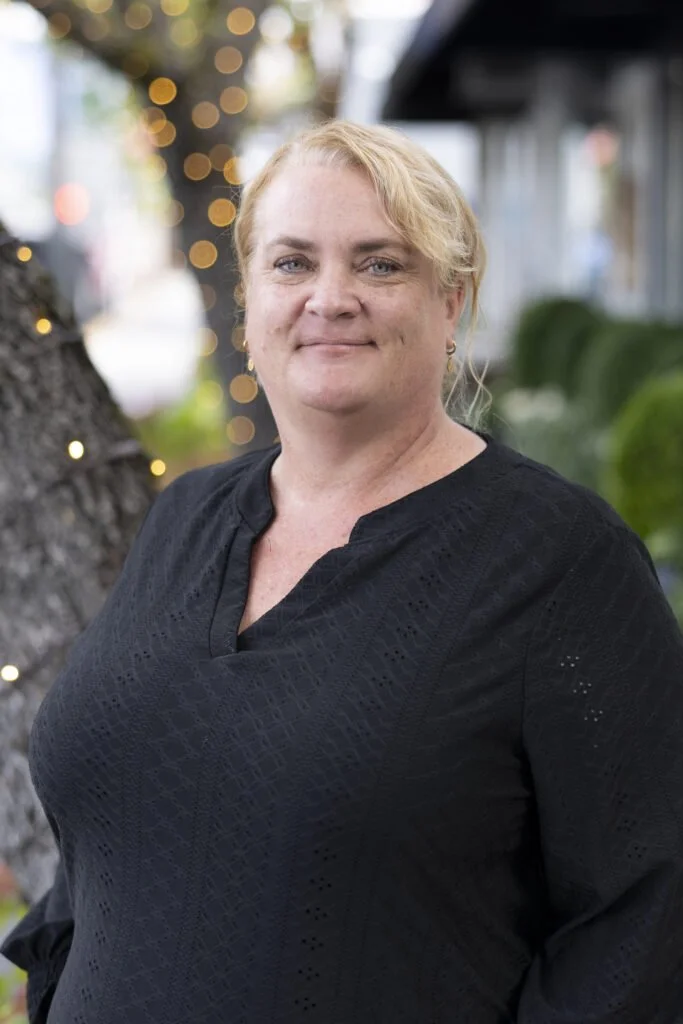 A woman with blond hair and light skin wears a black top, standing outdoors with blurred lights and foliage in the background.