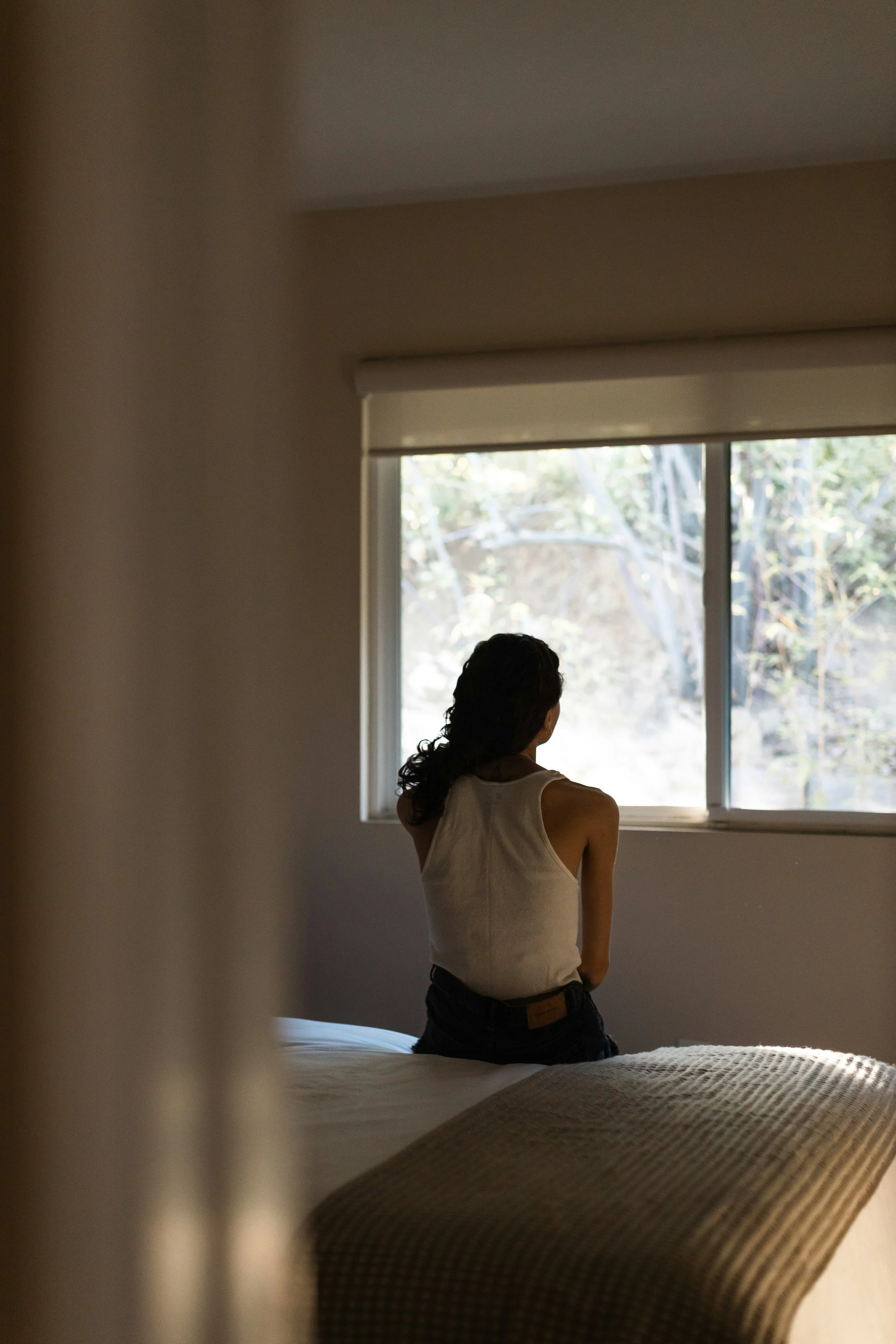 A woman with dark curly hair, wearing a sleeveless beige top and dark pants, stands by a window with trees outside, in a warmly lit room.
