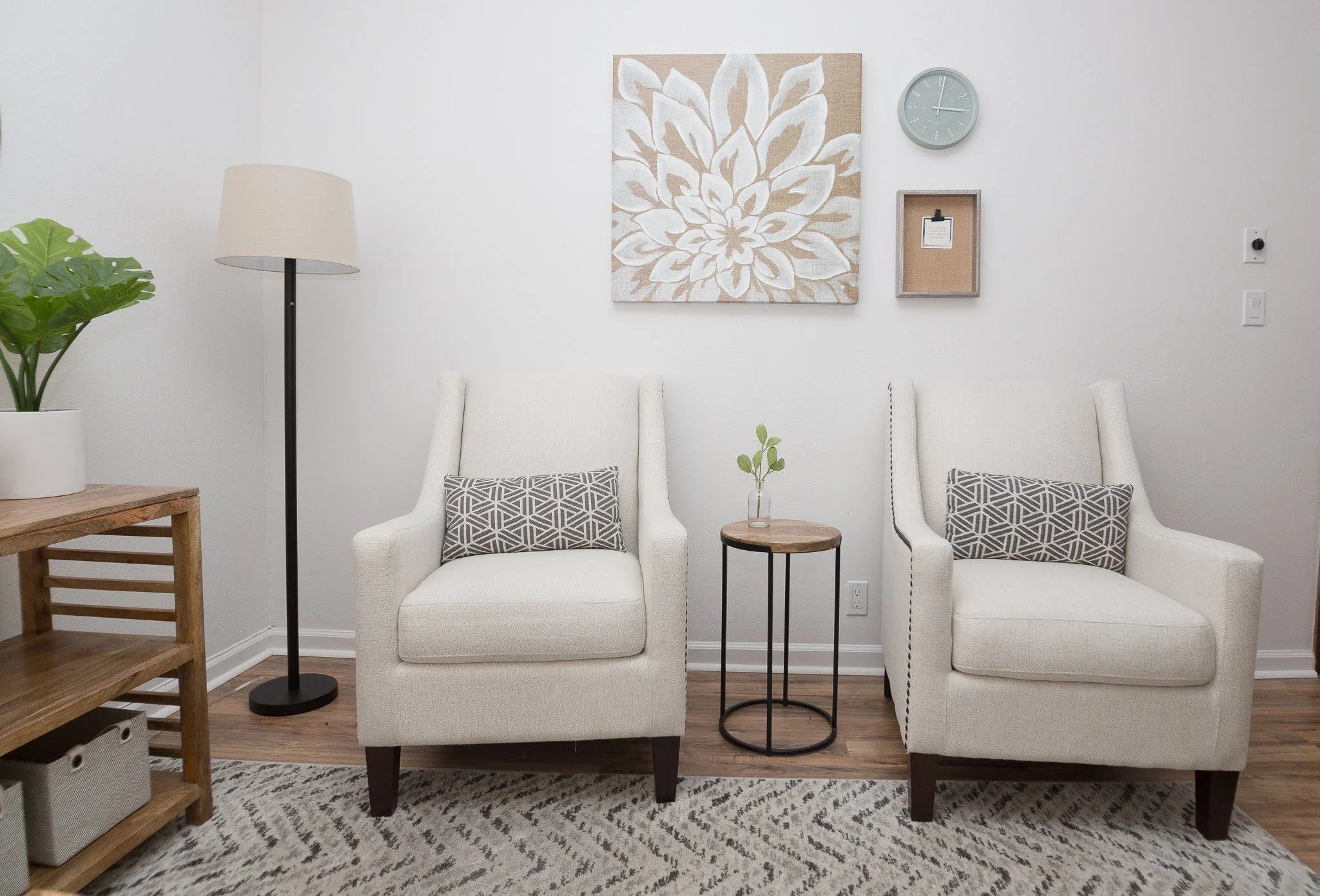 Living room with two white armchairs, small round side table, potted plant, wall art, and wall clock.
