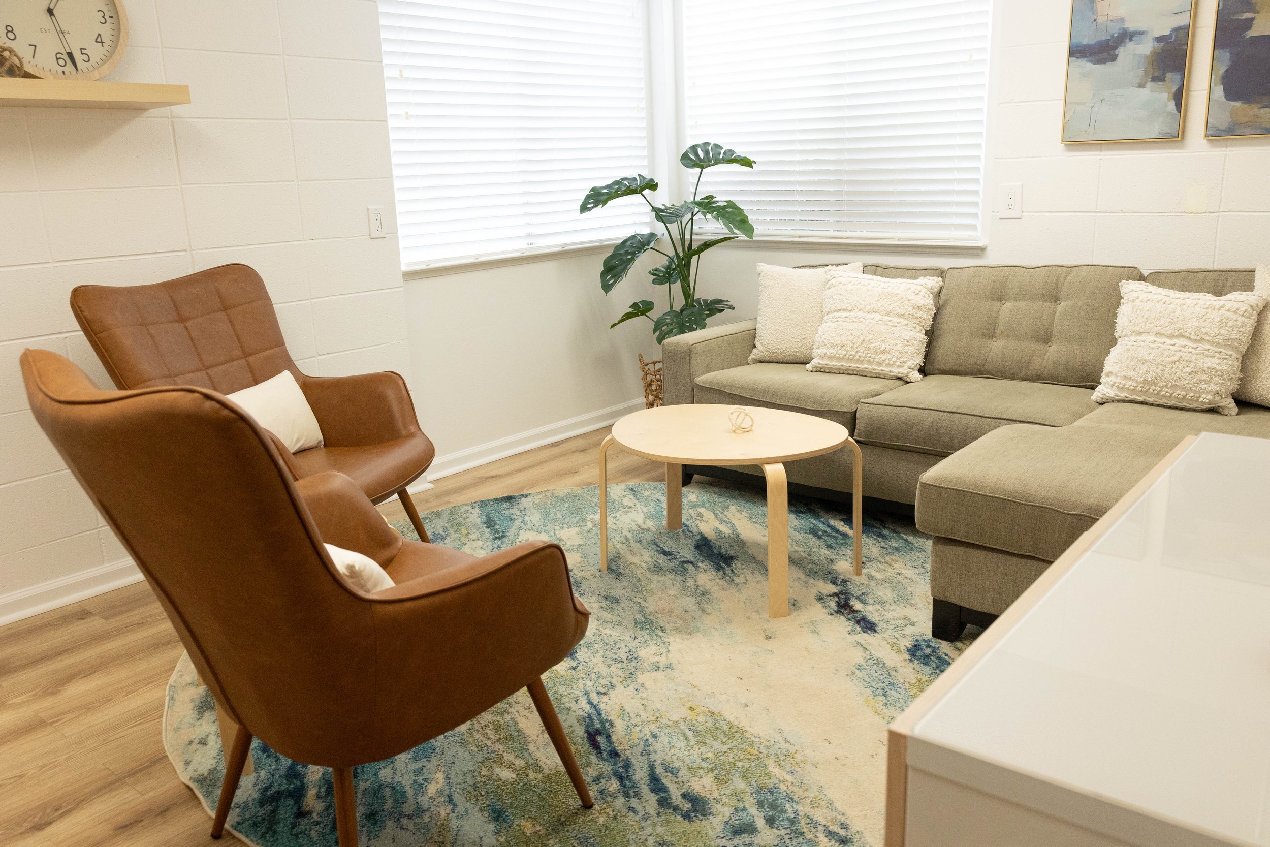 A cozy living room with a tan armchair, a gray sectional sofa with textured pillows, a round coffee table, a tall plant, framed artwork on the wall, and a large window with horizontal blinds.